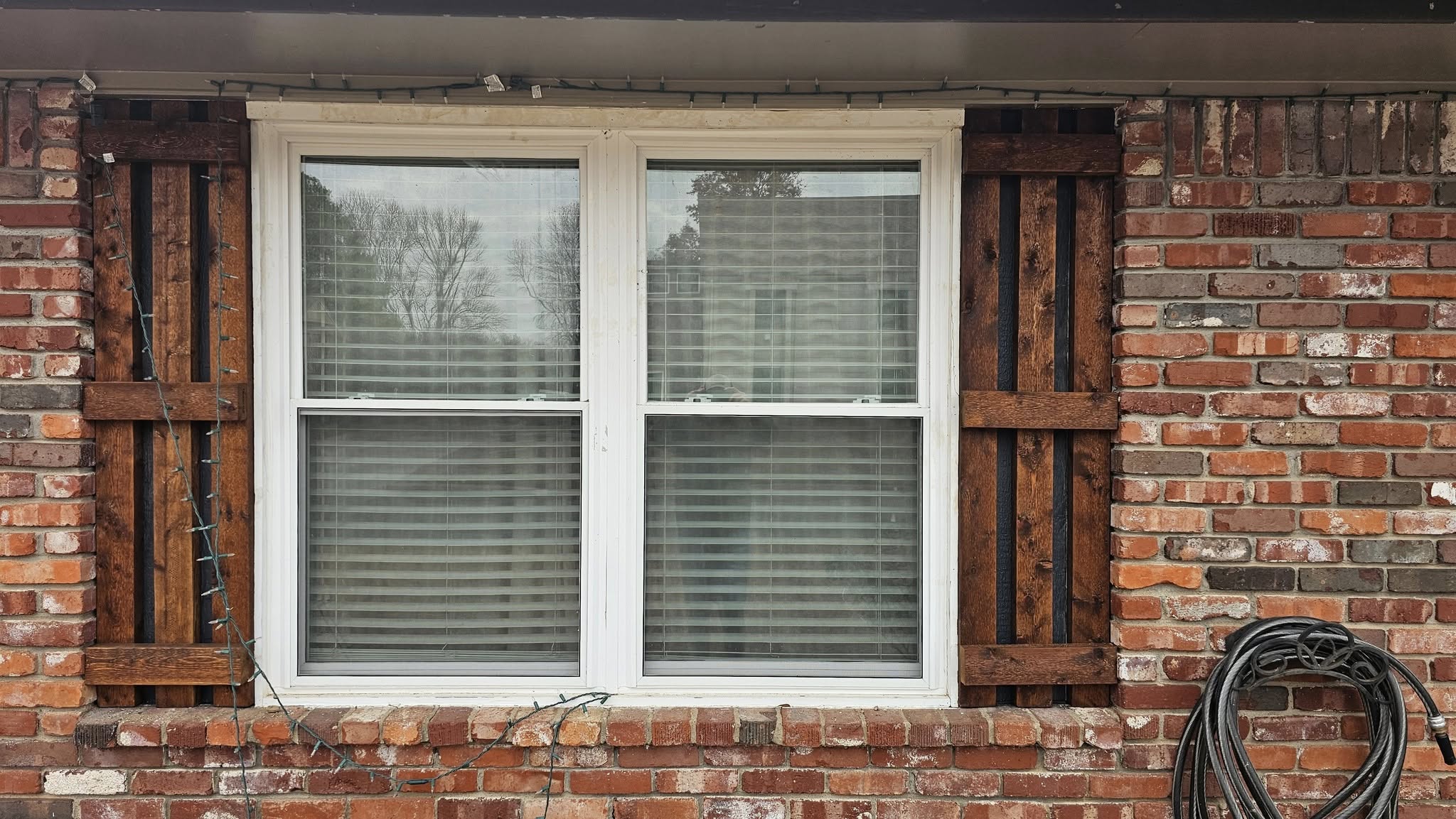 A window with wooden shutters and Christmas lights on a brick house.