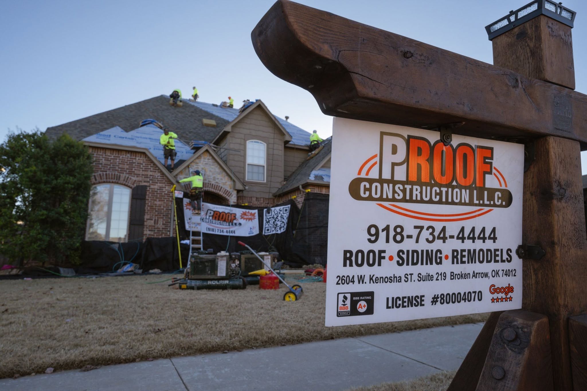 Roofers work on a house while a sign for Proof Construction L.L.C. stands nearby.
