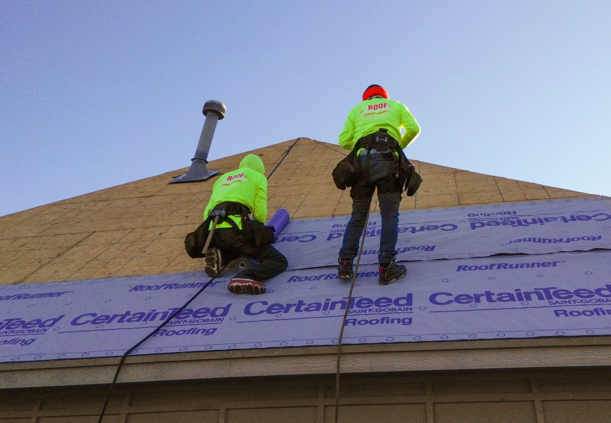 Two roofers in neon shirts work on a roof, installing underlayment.