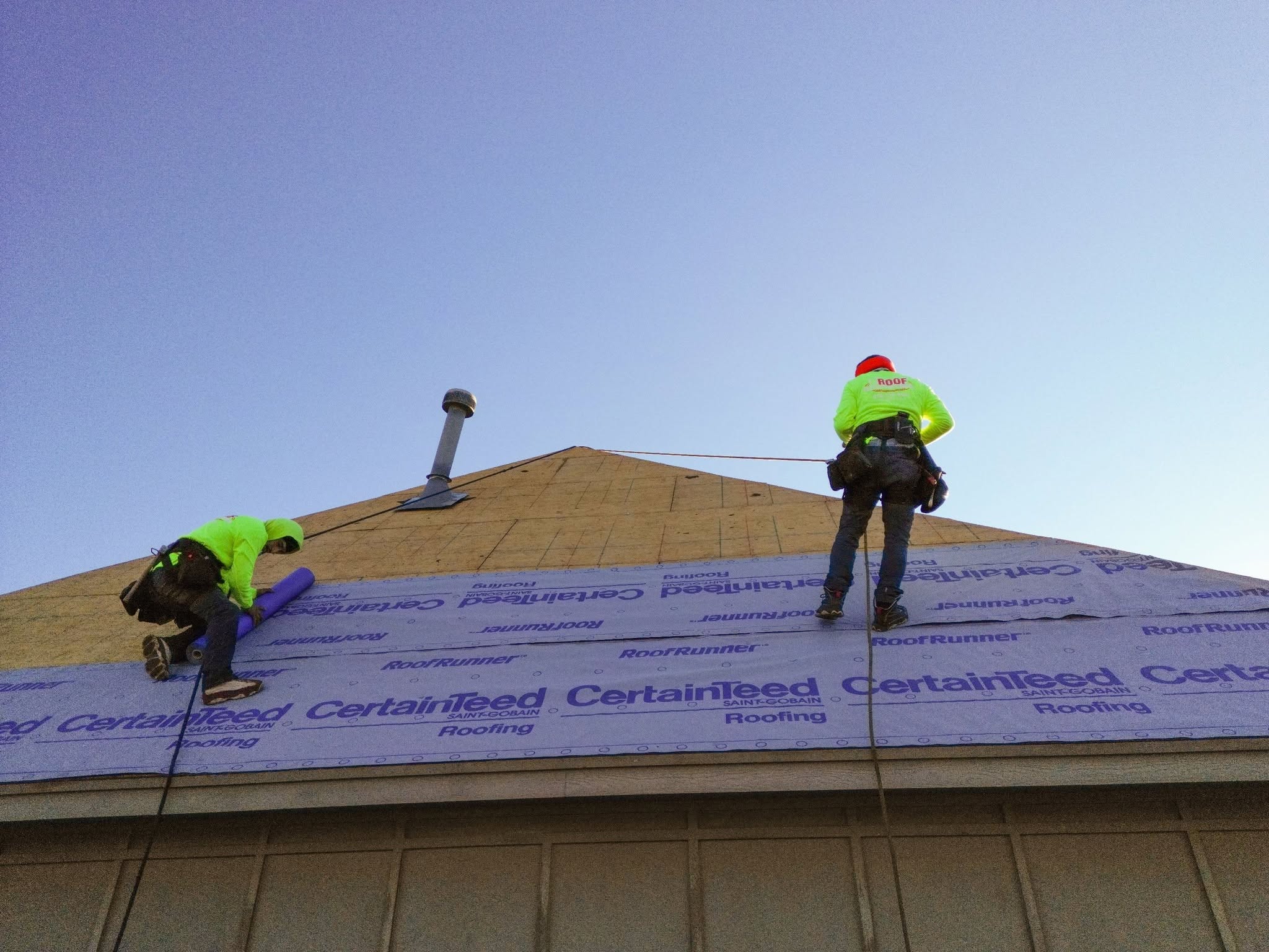 Two roofers in neon vests work on a house roof, installing underlayment.