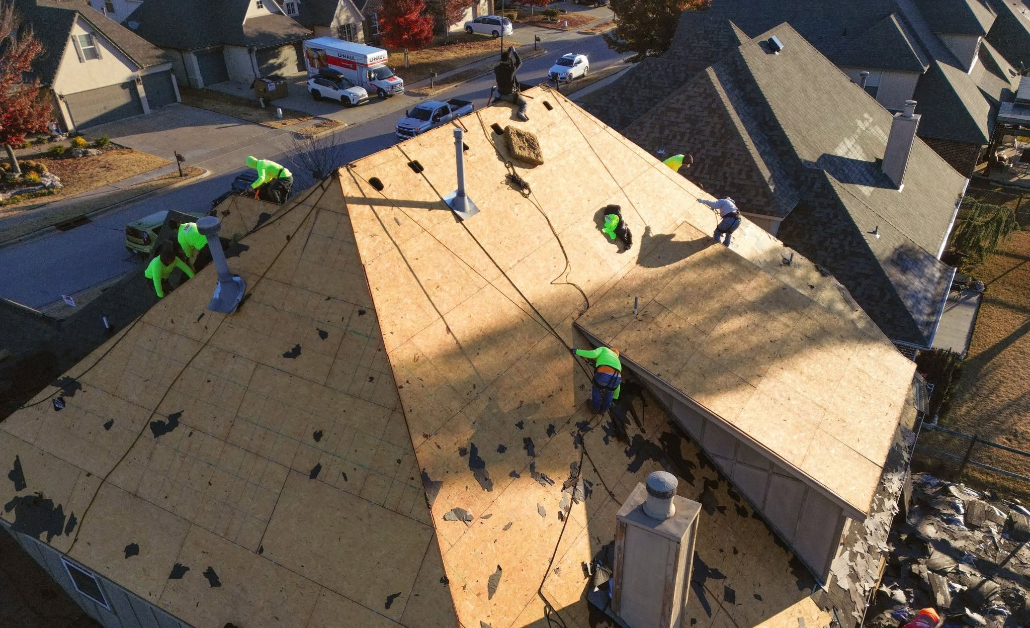 Roofers work on a house roof, with U-Haul trucks and cars visible in the background.