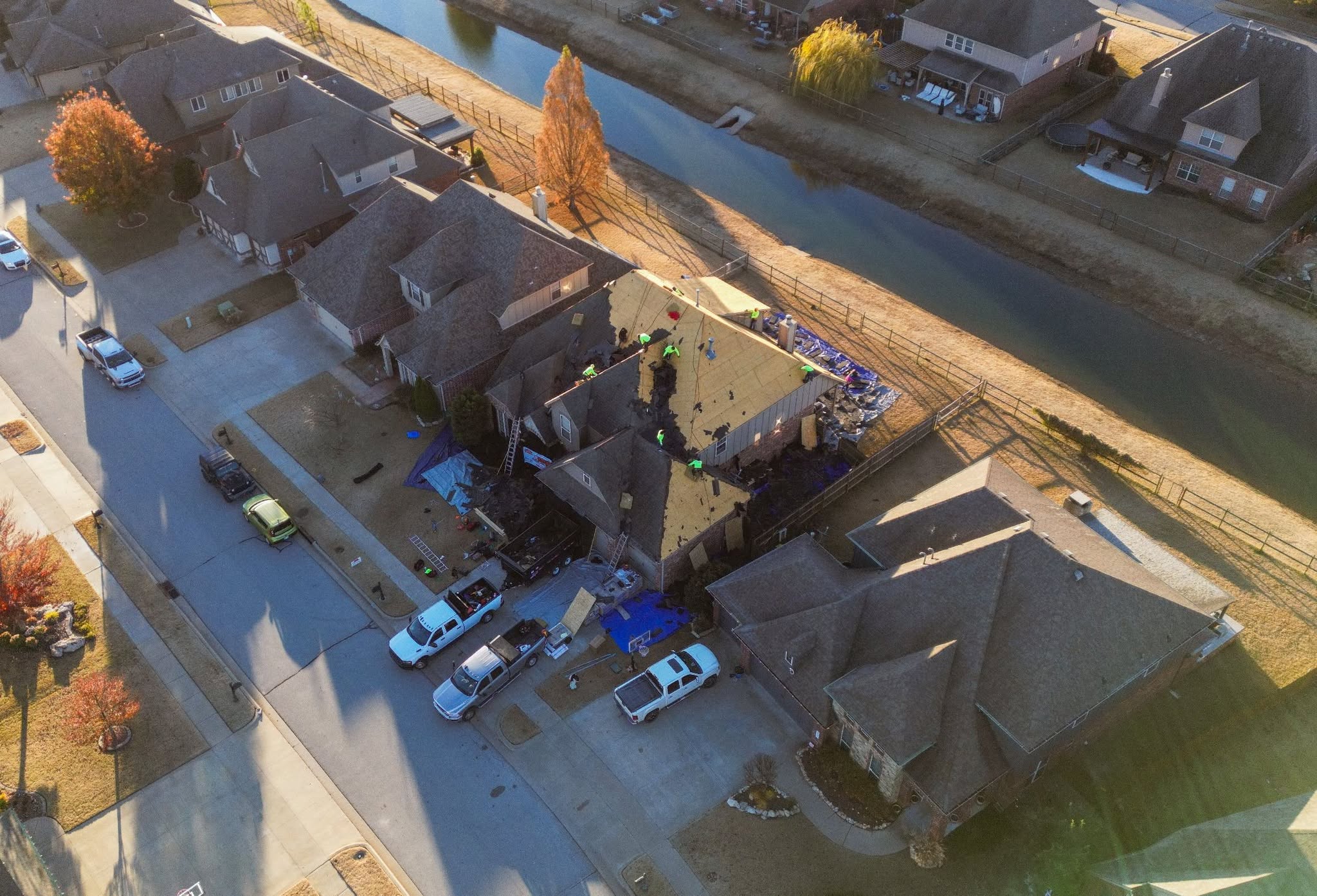 Roofers work on a damaged house with vehicles and debris in a suburban neighborhood.