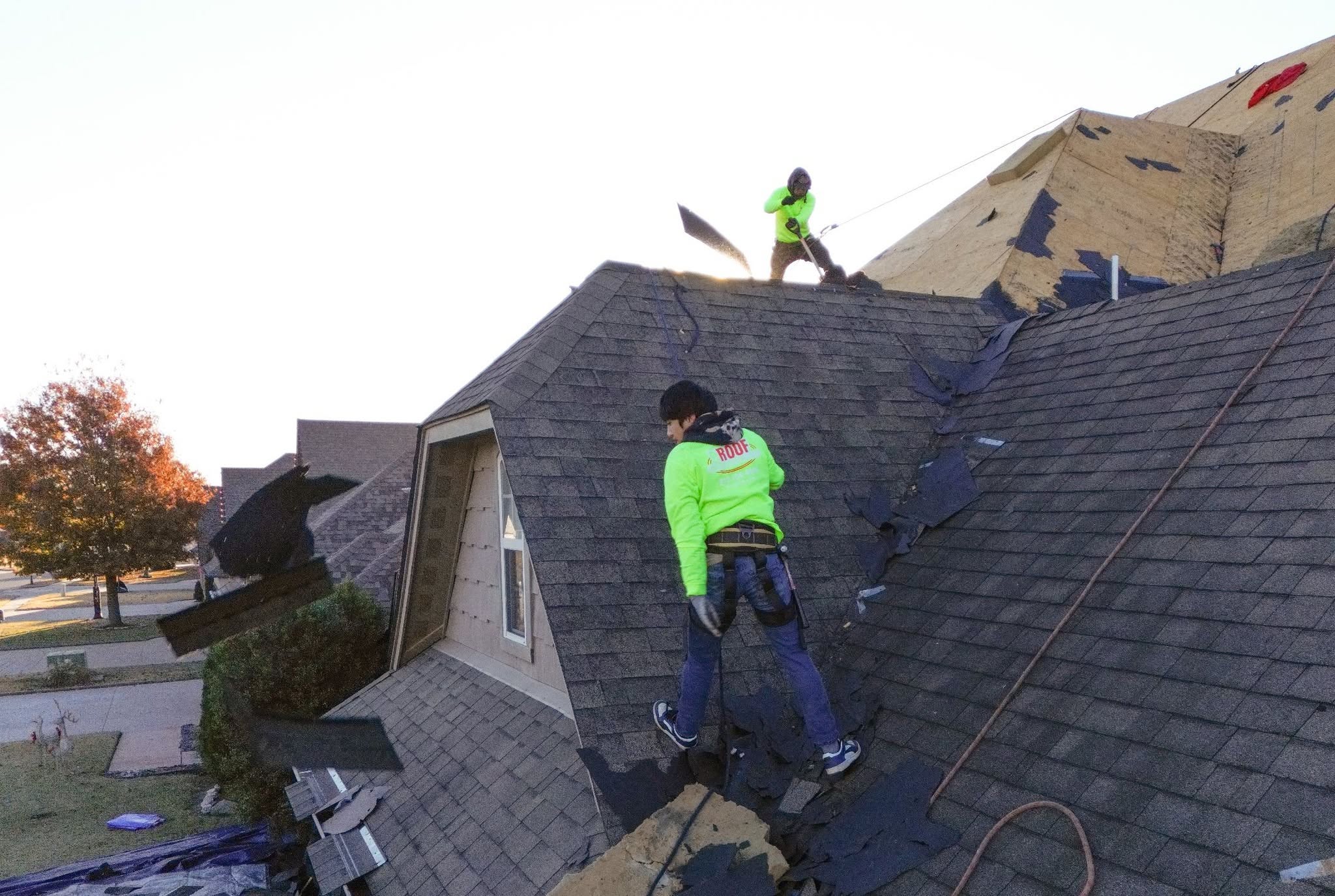Two roofers in neon shirts work on a damaged roof in a suburban neighborhood.