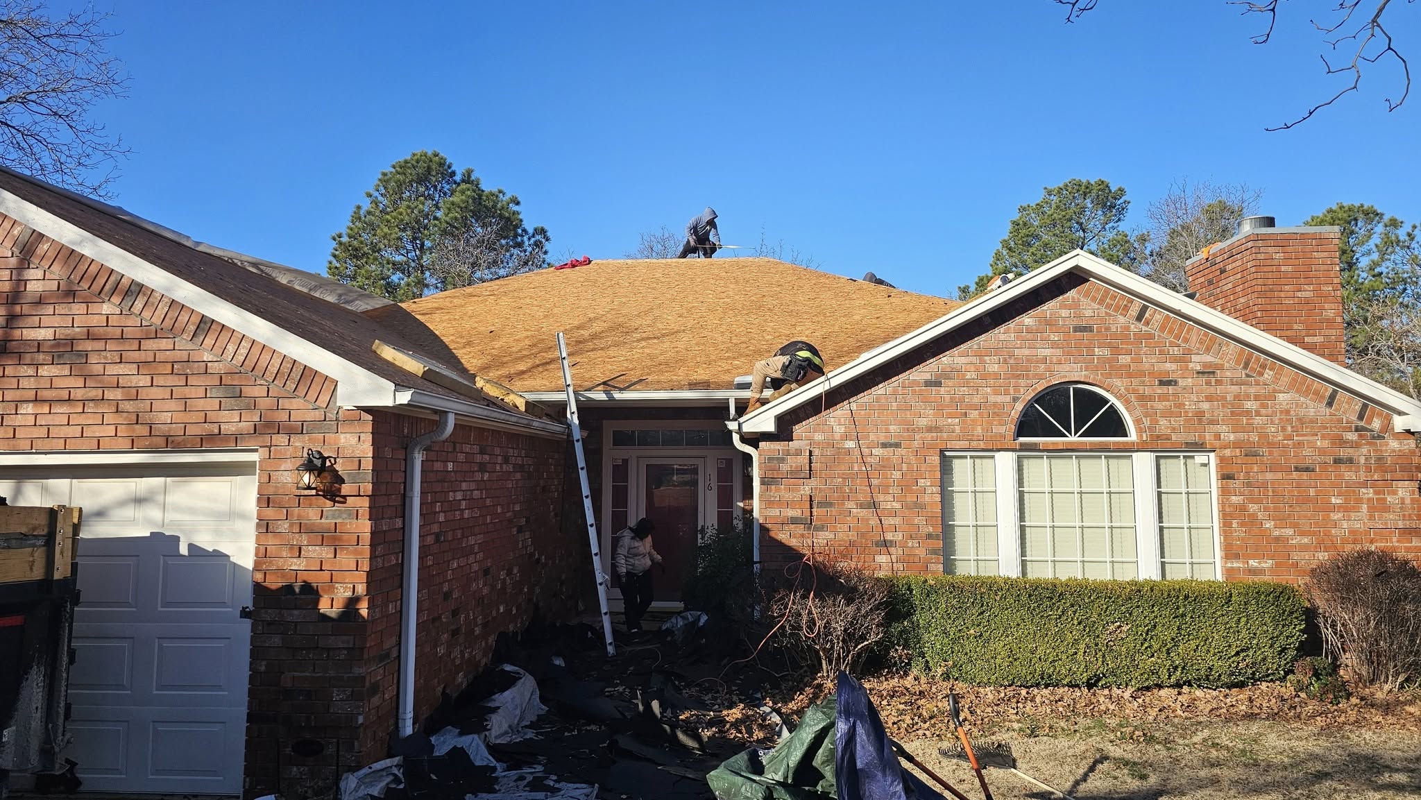 Roofers work on a house's roof, with a ladder, debris, and tools visible.
