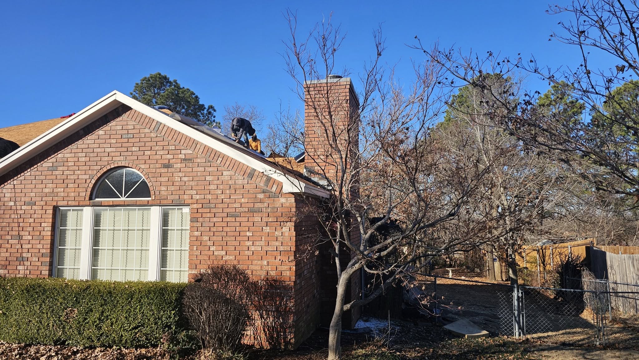 Two roofers work on a brick house with a chimney on a sunny day.