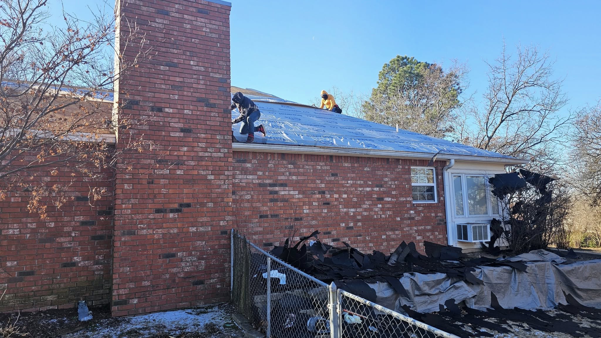 Two roofers work on a house with a brick chimney, while debris is piled on a tarp below.