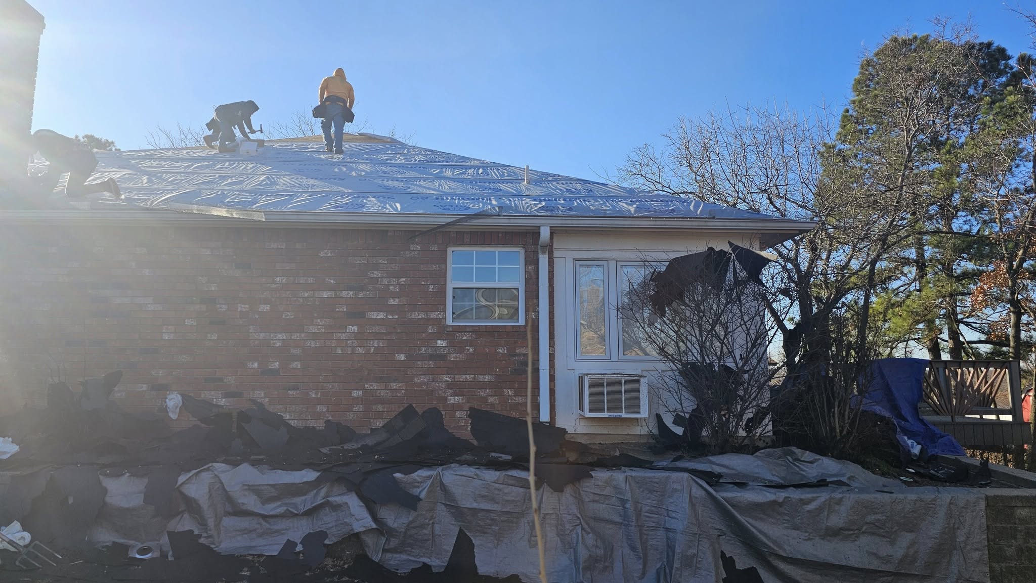 Roofers work on a house covered in protective sheeting, with debris piled below.