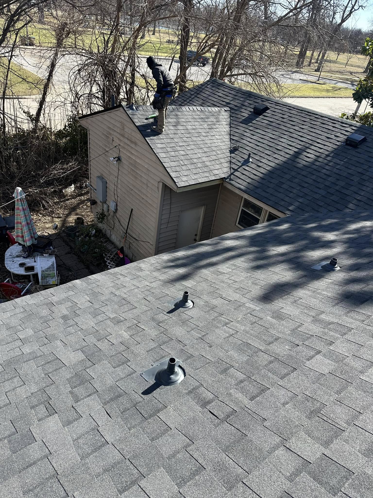 A roofer works on a grey shingled roof of a house with trees in the background.