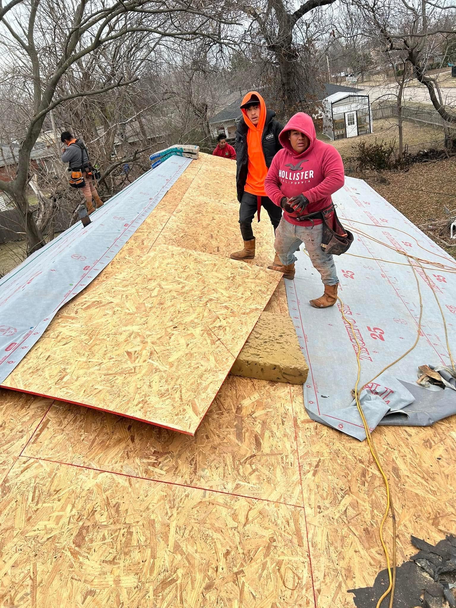Two construction workers are on a roof, installing plywood and underlayment.