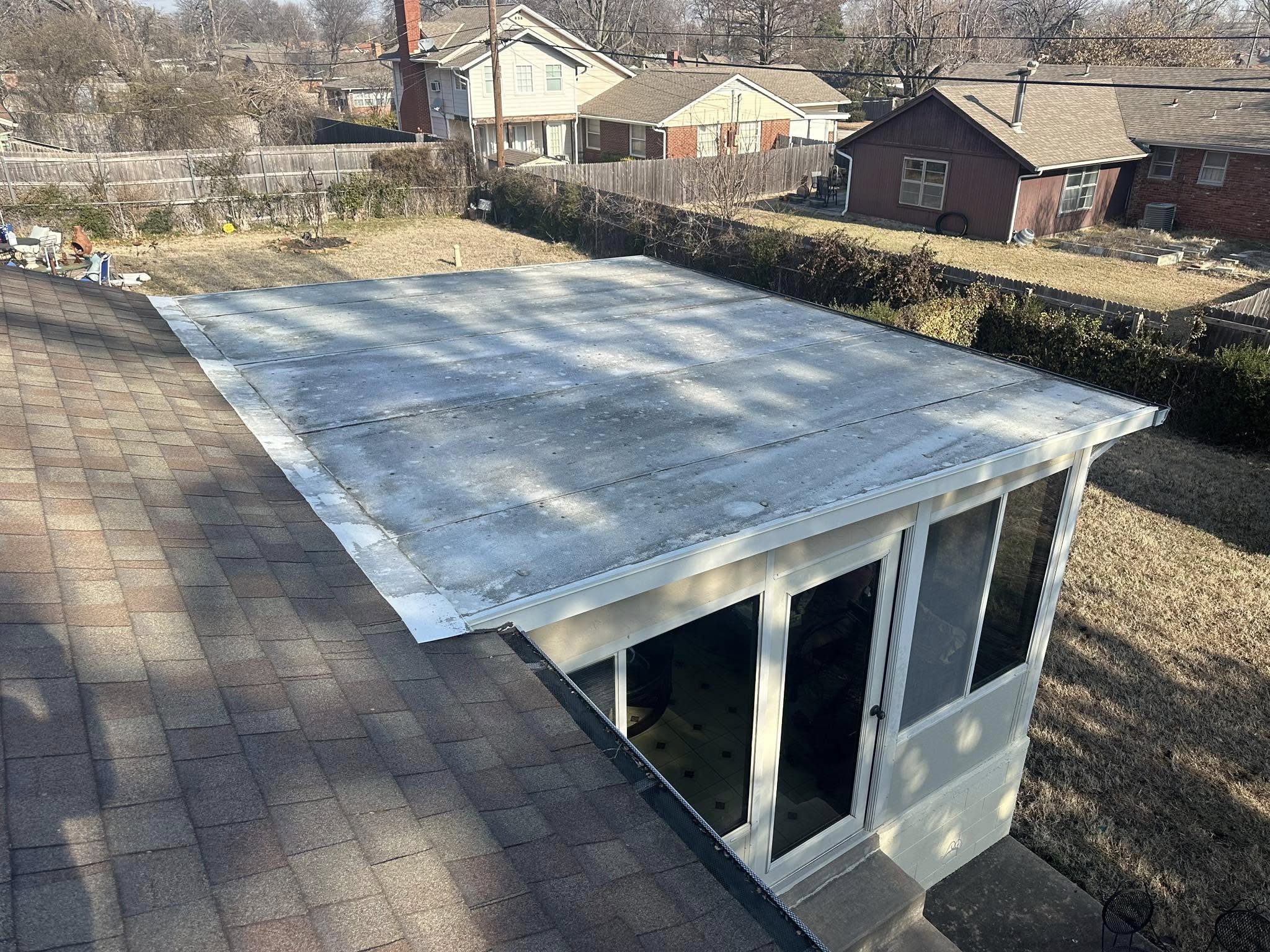 A flat roof extension with a shingled roof is shown on a sunny day in a suburban backyard.