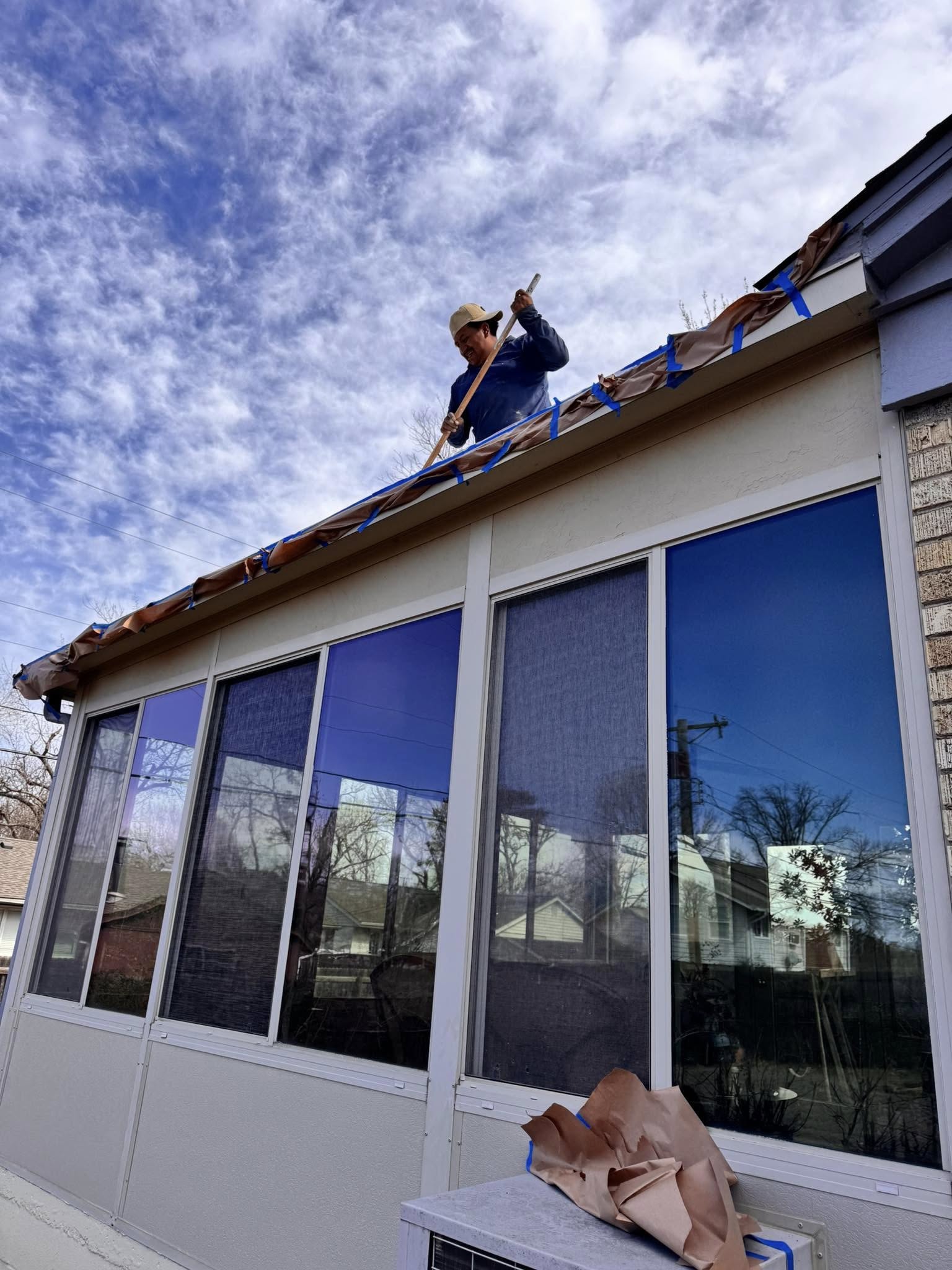 A man on a roof is painting the eaves of a sunroom with blue sky above.