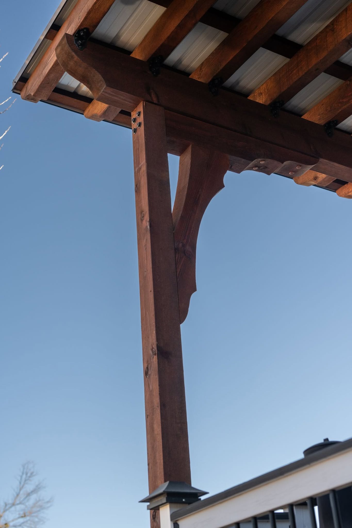 A wooden pergola with a metal roof is supported by a post against a clear blue sky.