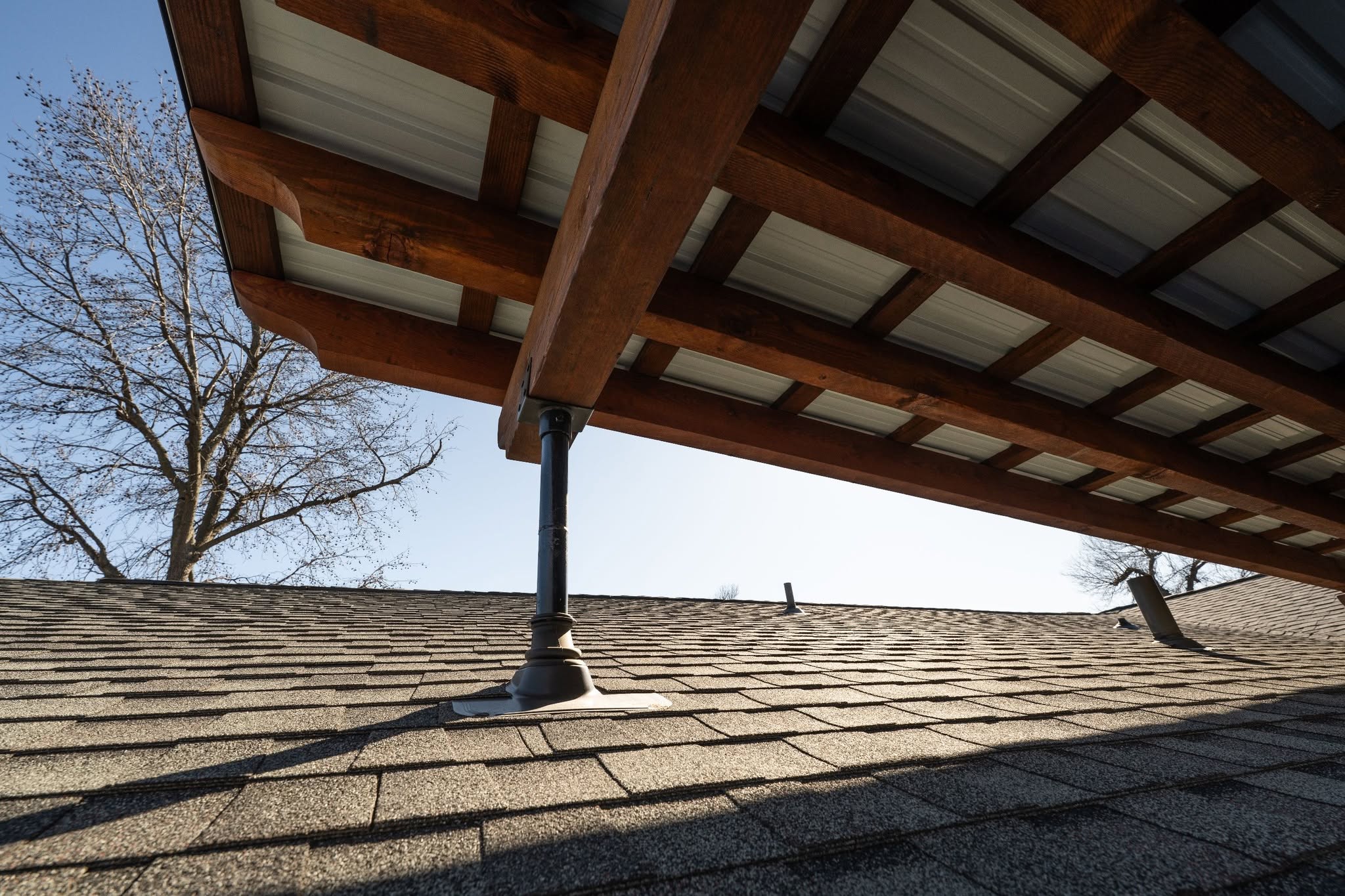 A wooden roof structure is supported by a metal post on a shingled roof.
