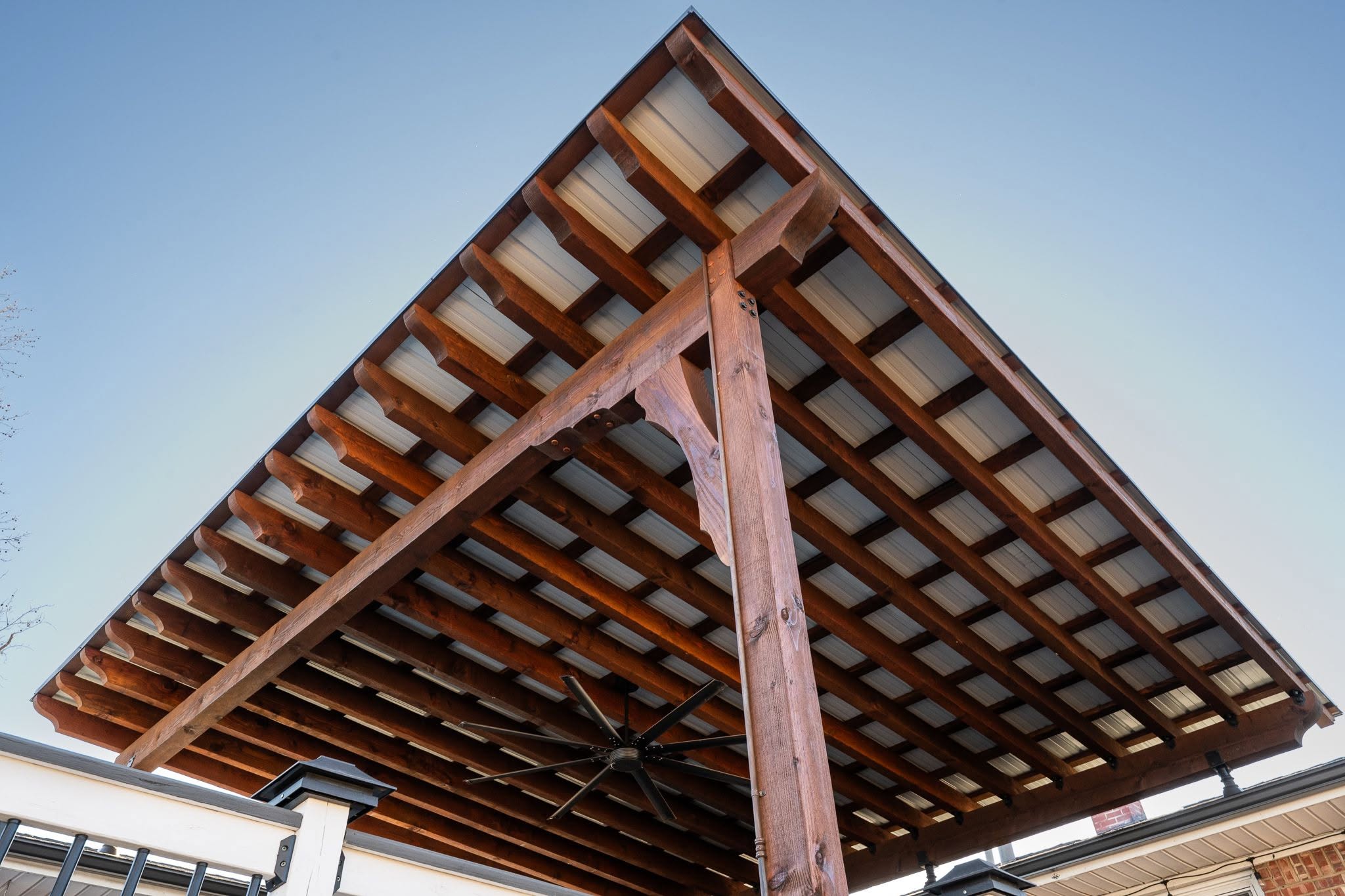 A wooden pergola with a metal roof and a fan is shown outdoors.