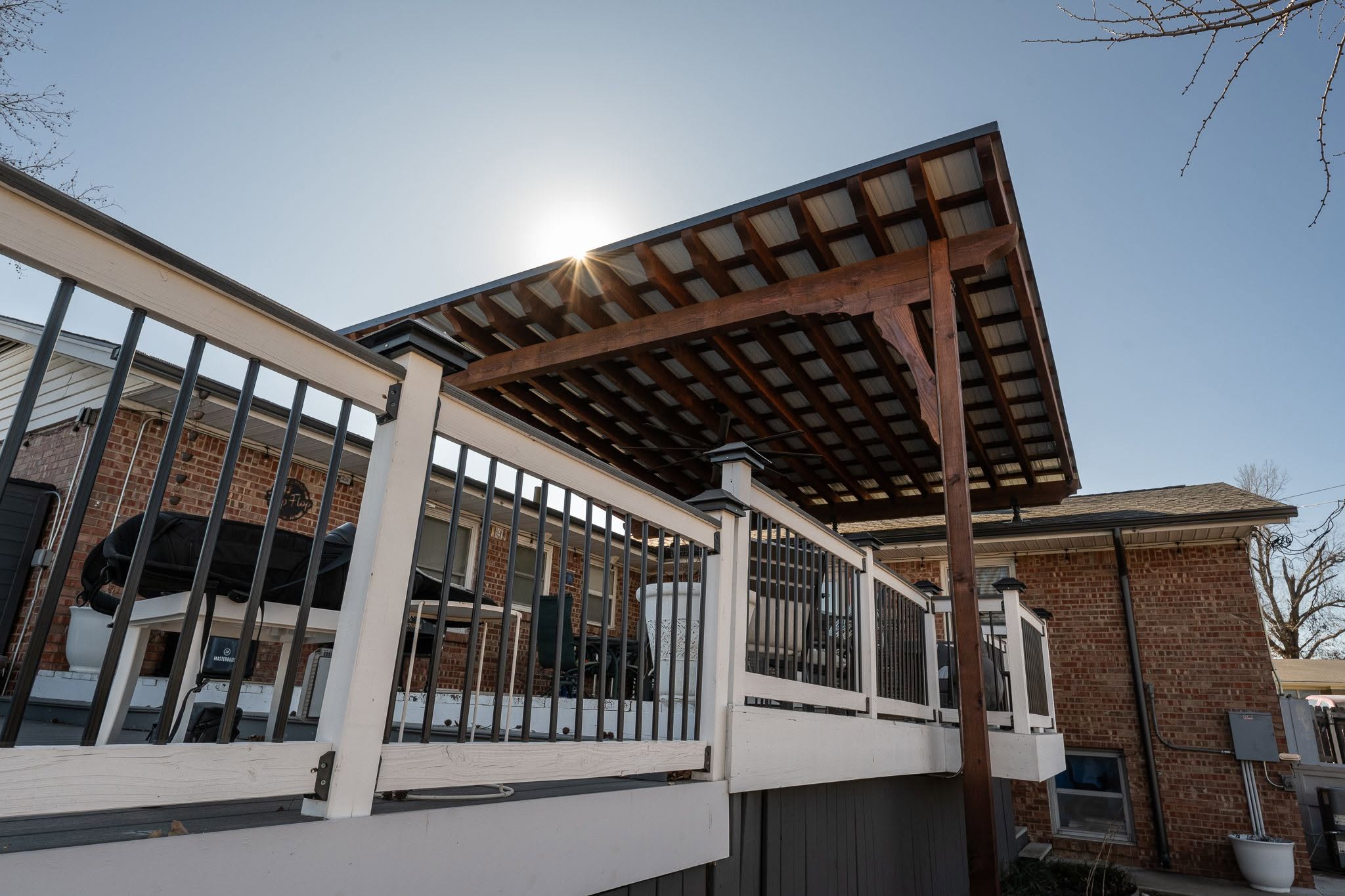 A covered deck with a wooden pergola overlooks a brick house and yard on a sunny day.