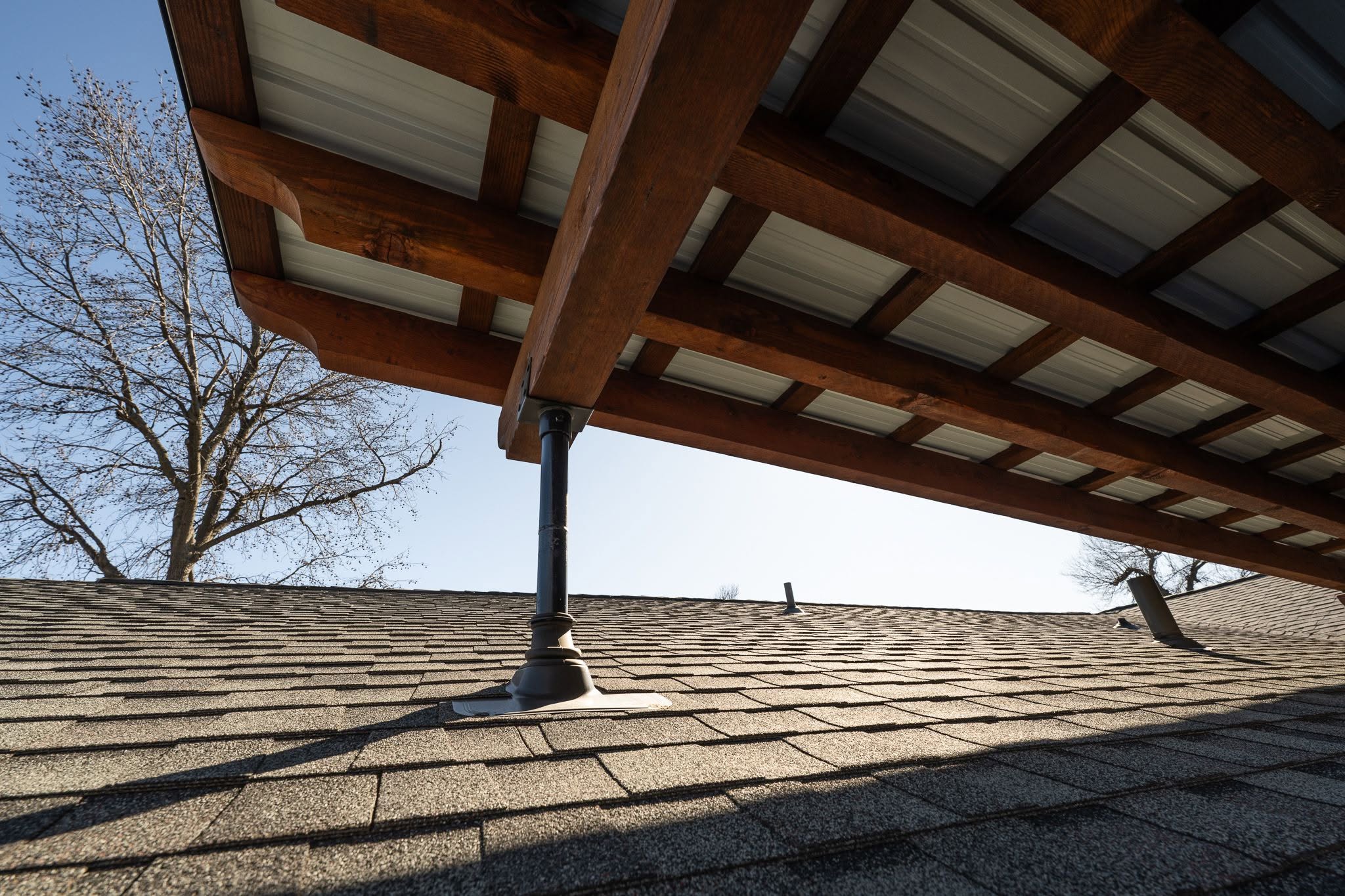 A wooden roof structure is supported by a metal post on a shingled roof.