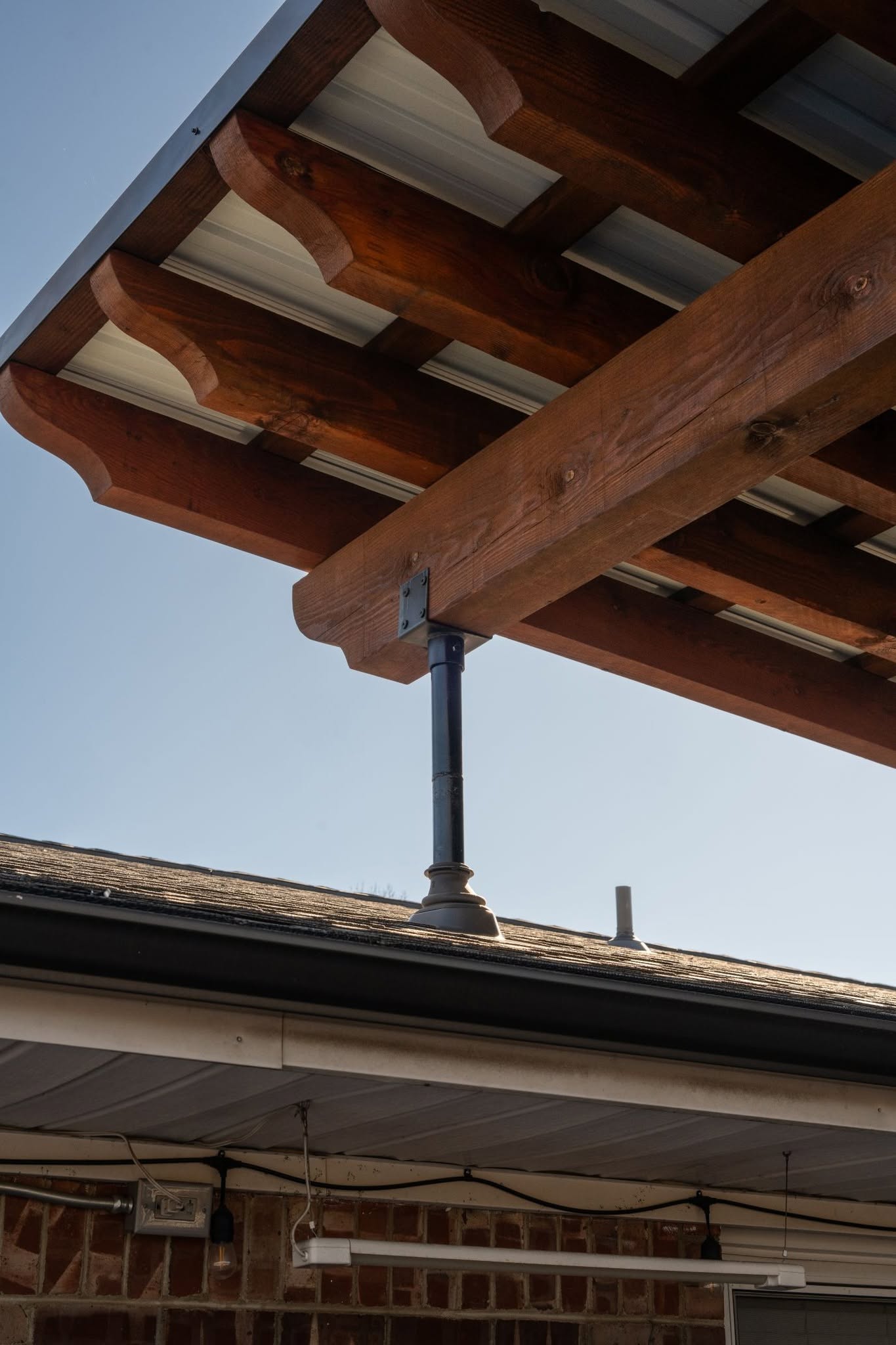A wooden pergola with a metal roof is supported by a black metal post over a shingled roof.