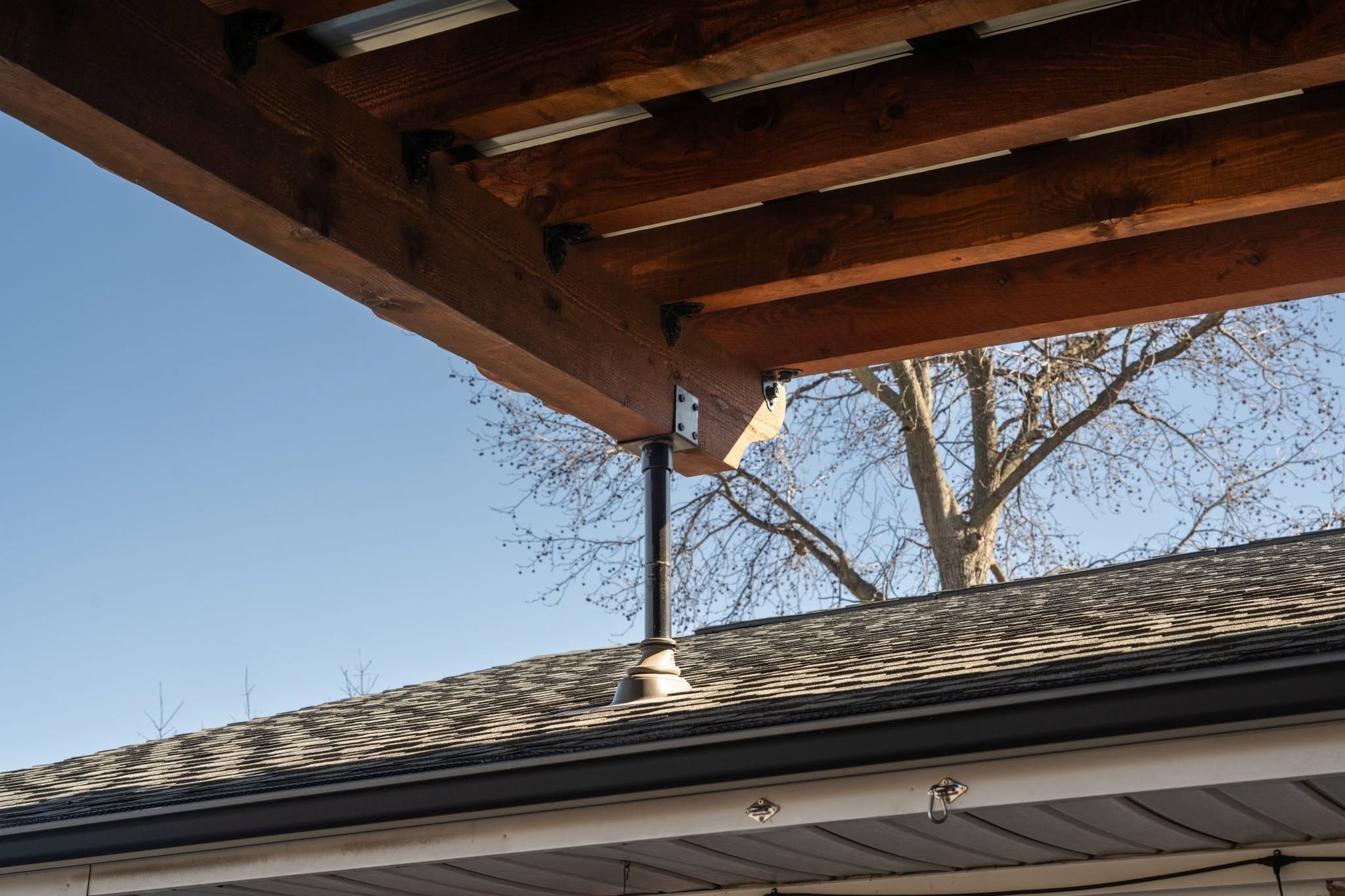A wooden pergola is supported by a metal post above a shingled roof.