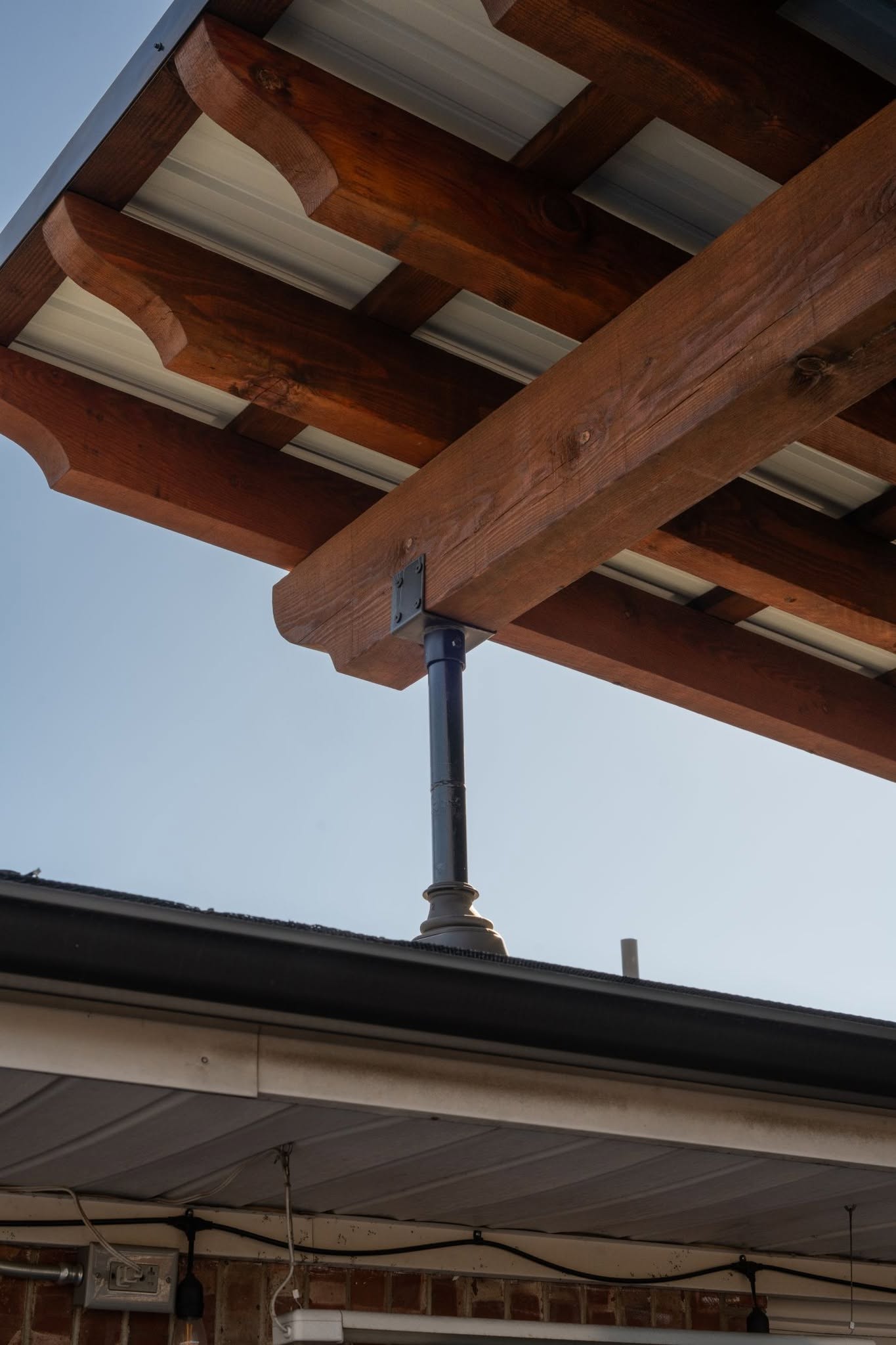 A wooden pergola with a metal roof is supported by a black metal post over a house's gutter.