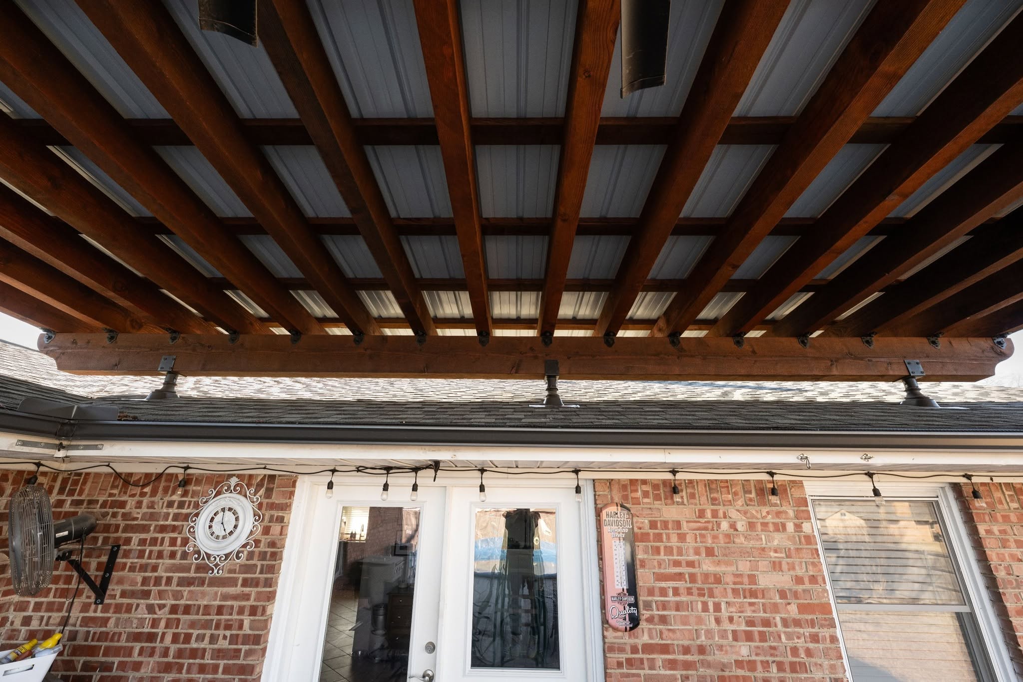 A wooden pergola covers a brick patio with a clock and fan.
