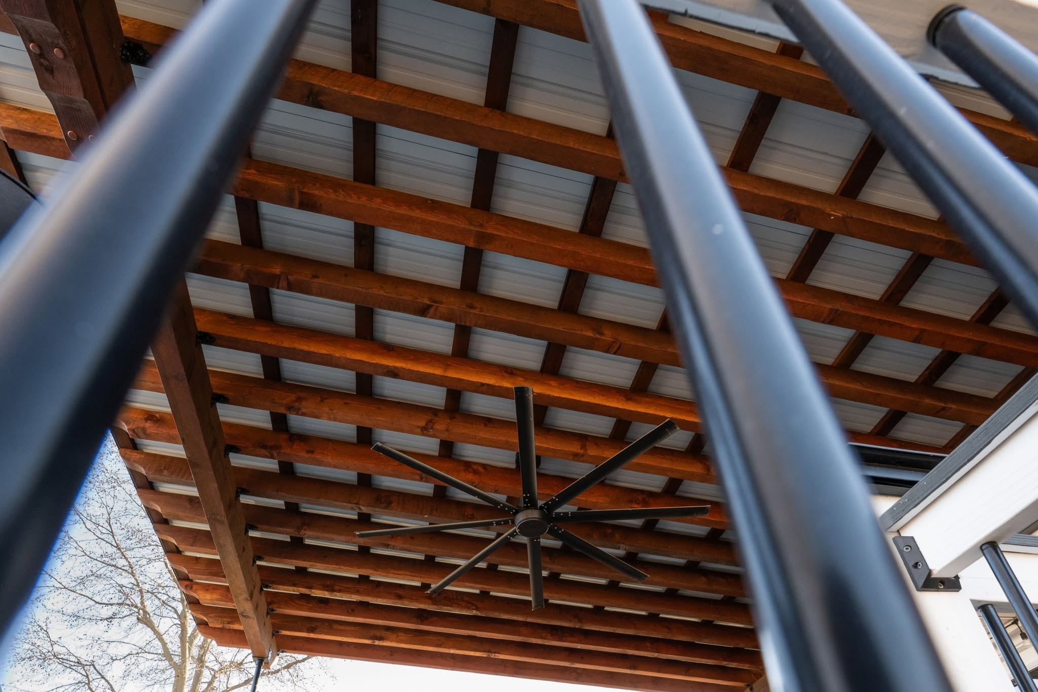 A large black ceiling fan is mounted on a wooden beam ceiling, viewed through dark vertical bars.
