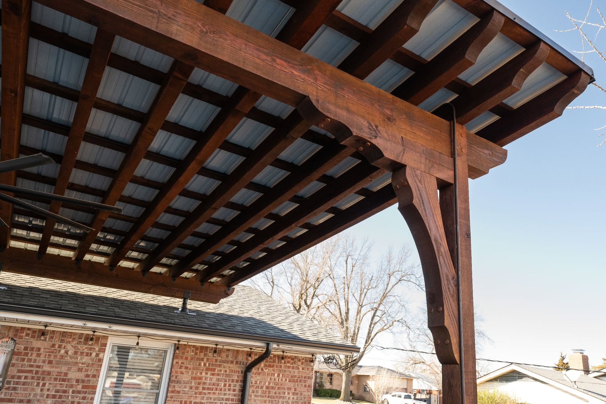 A wooden pergola with a metal roof is attached to a brick house.