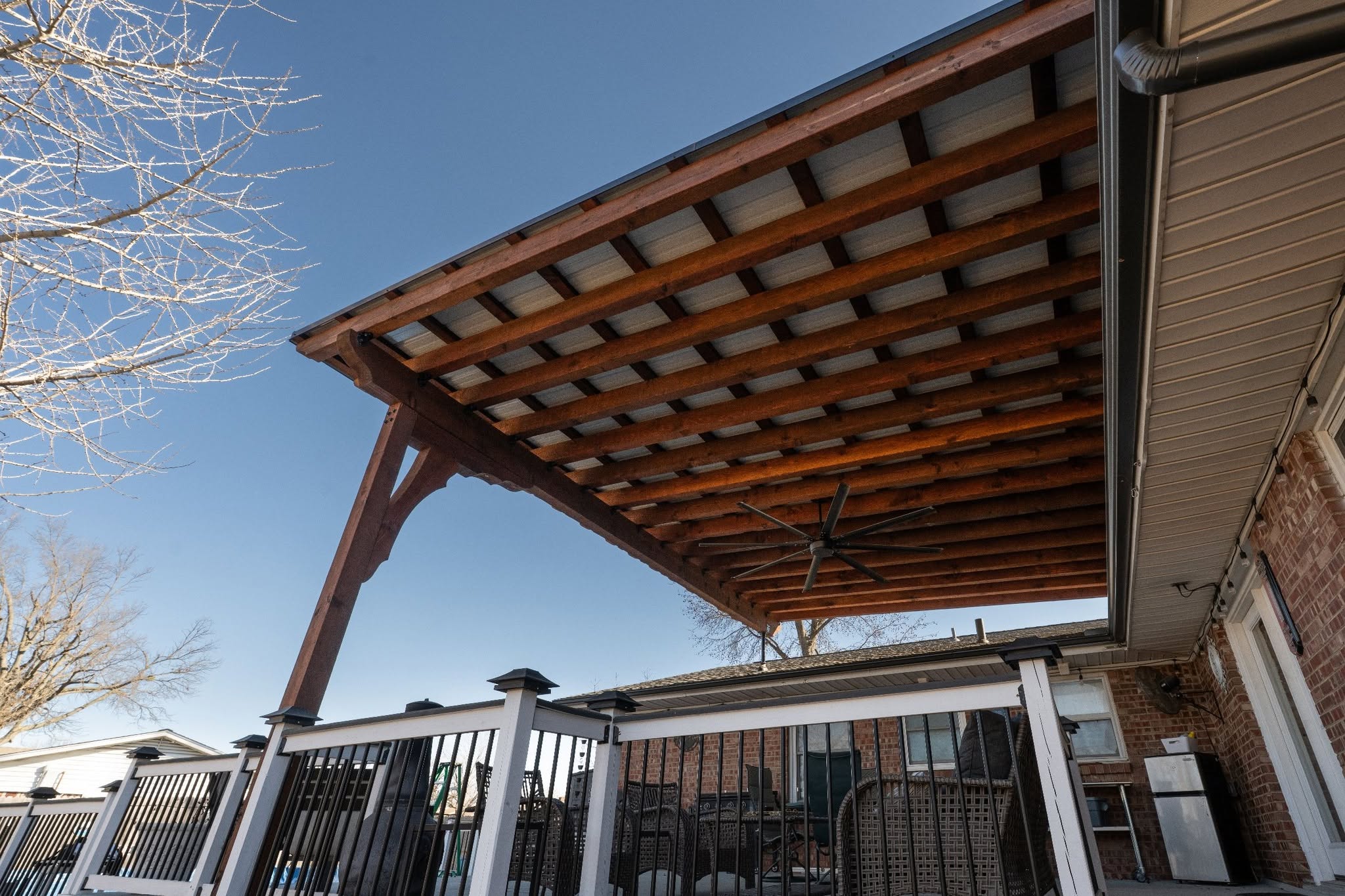A covered patio with a ceiling fan and outdoor furniture is shown on a sunny day.