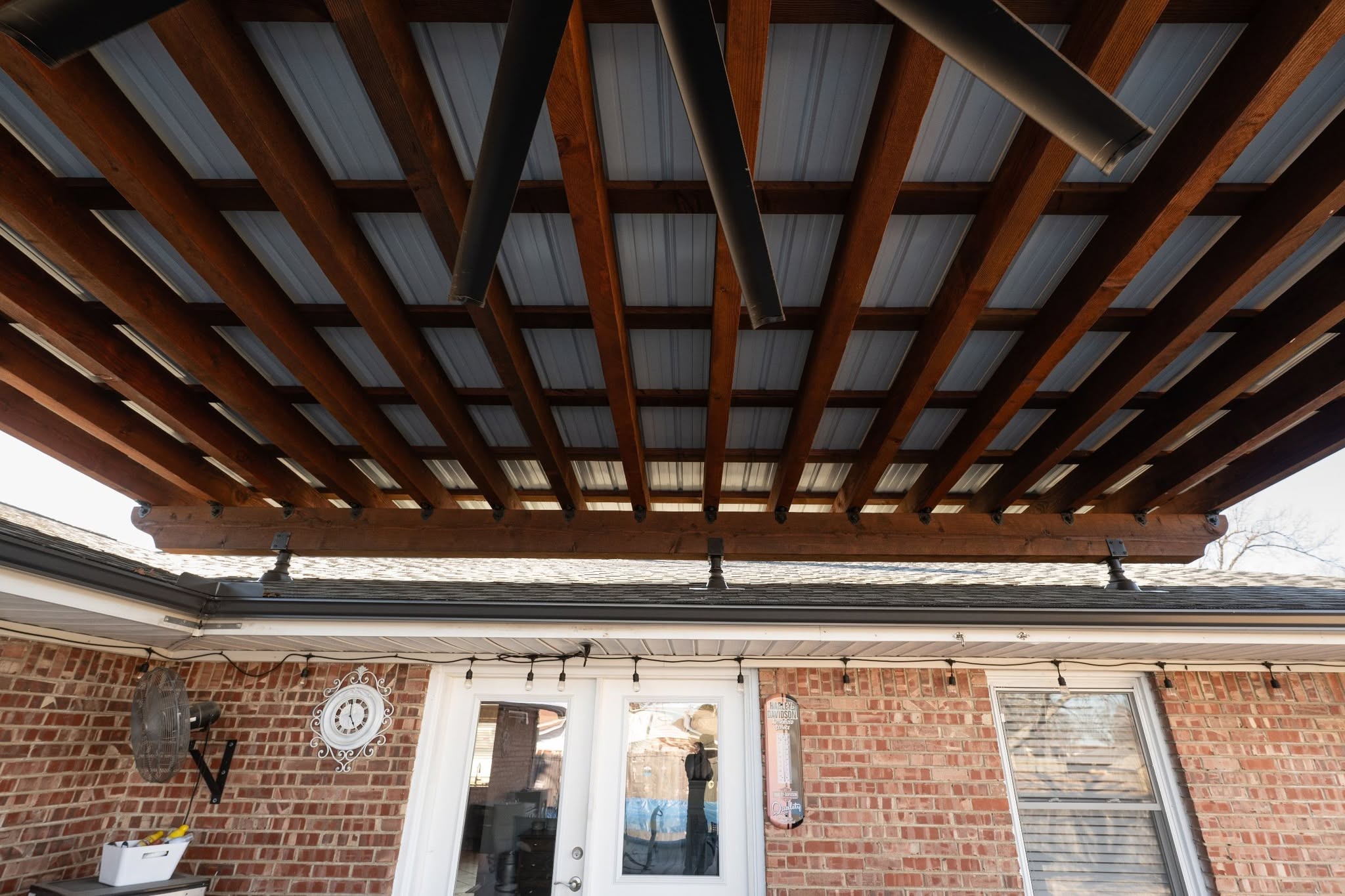 A covered patio with a wooden beam ceiling, metal roof, and brick wall with a fan and clock.