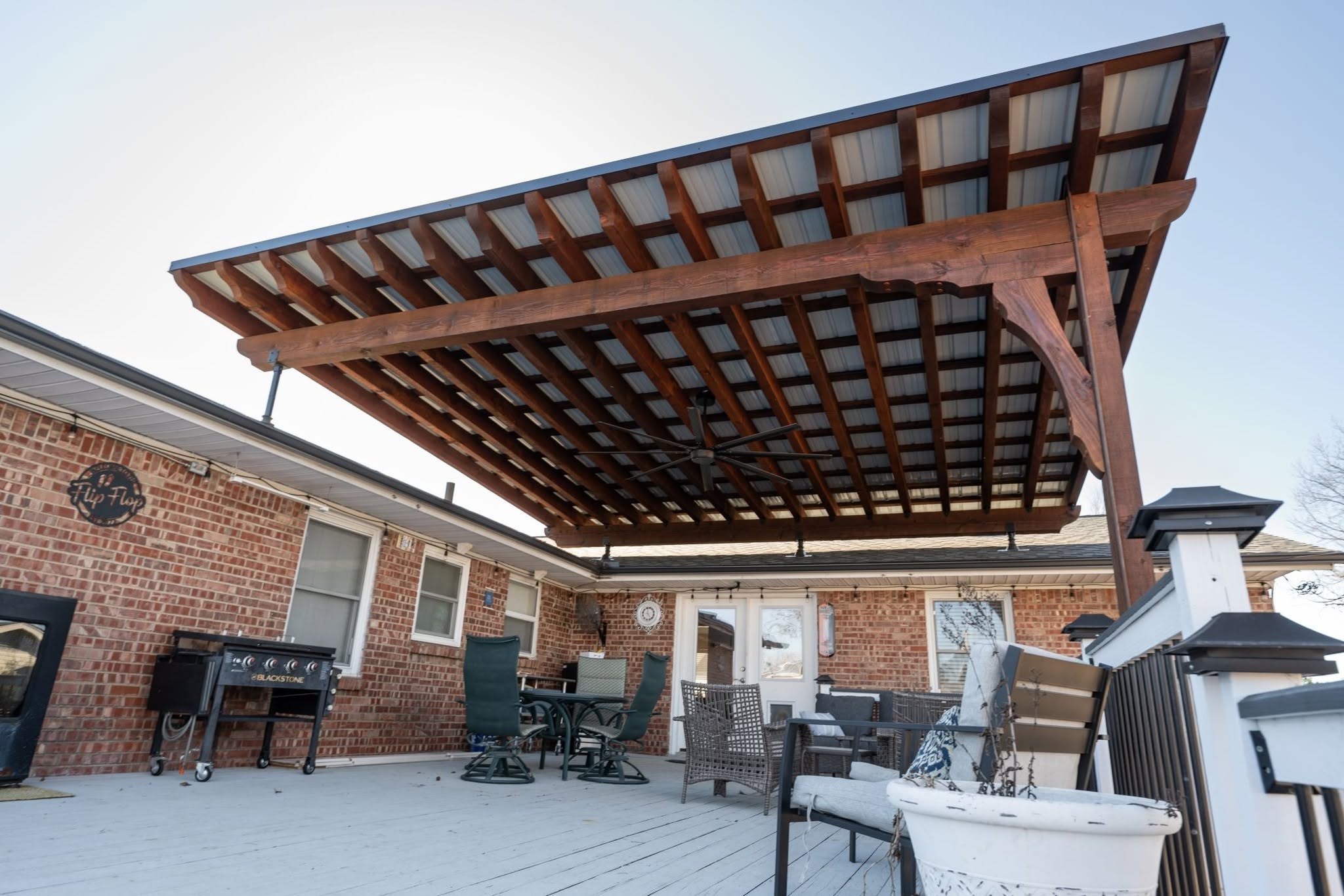 A covered patio with furniture and a grill is shown on a deck outside a brick house.