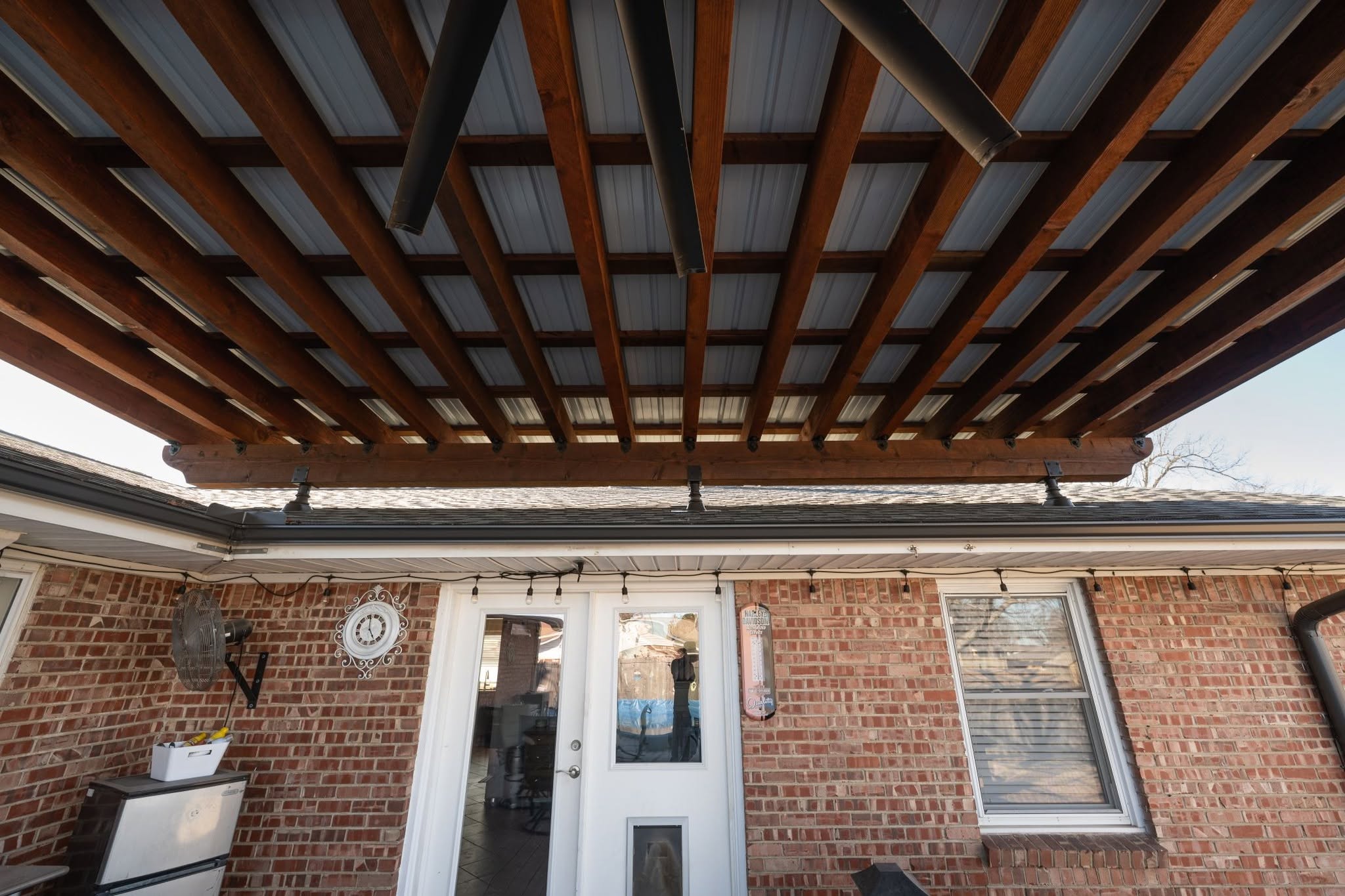 A covered patio with a brick house, featuring a fan, clock, and string lights.
