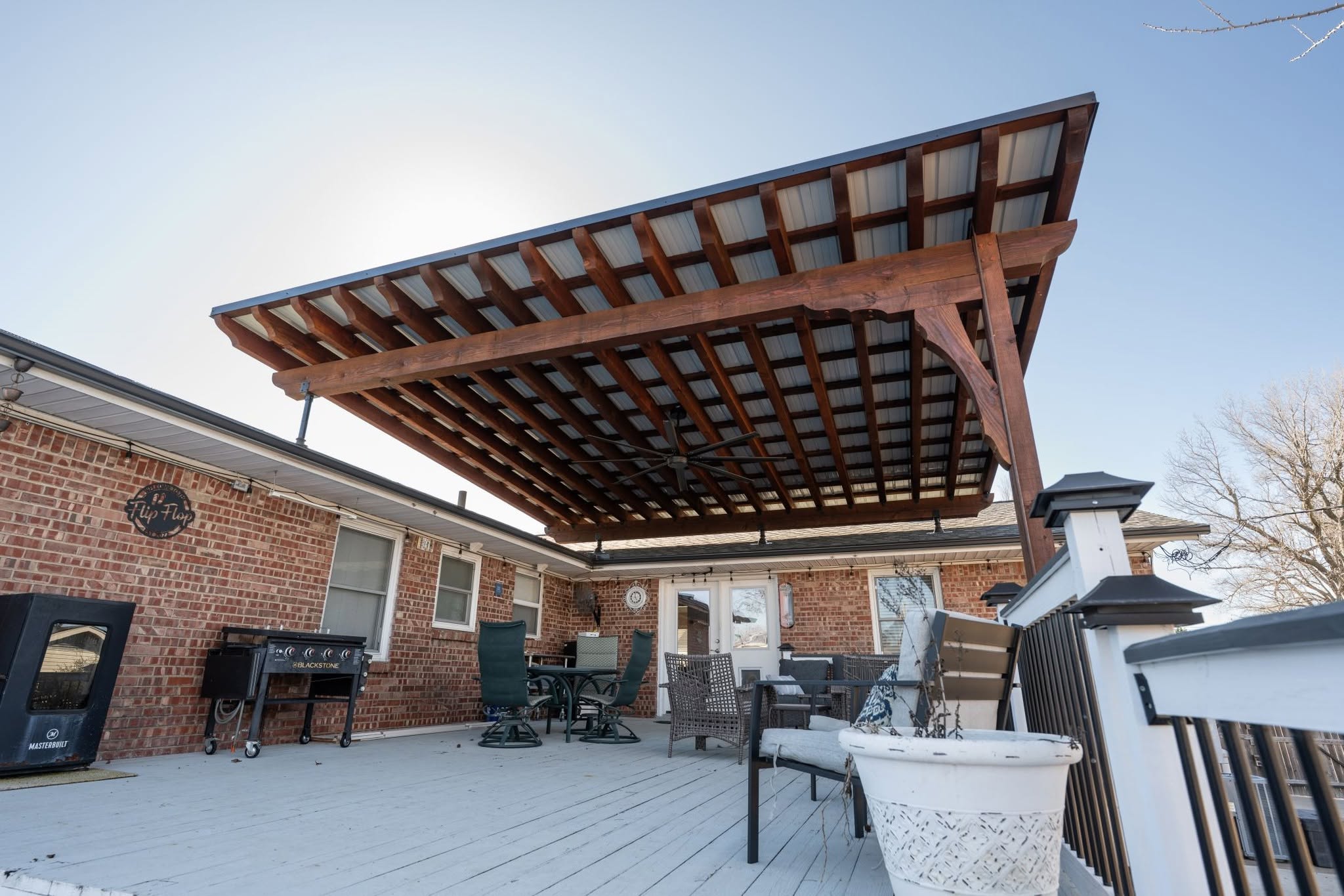 A covered patio with outdoor furniture and a grill on a wooden deck outside a brick house.