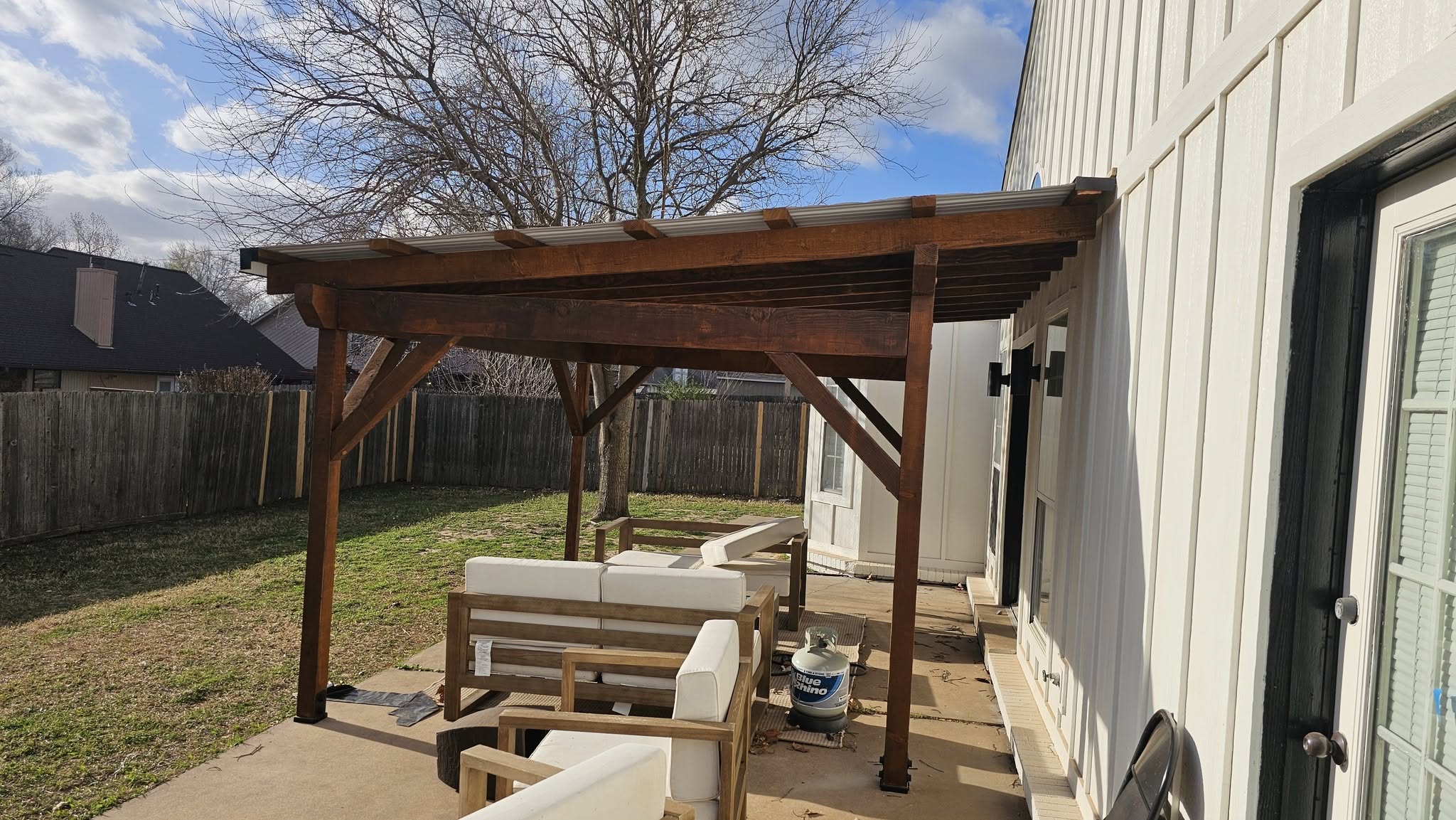 A wooden pergola covers outdoor furniture and a propane tank in a backyard.