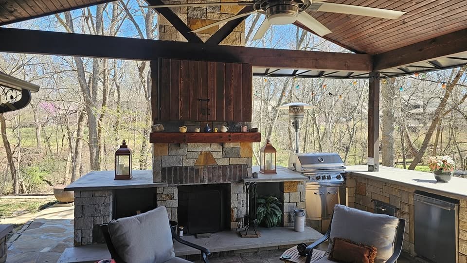 An outdoor kitchen with a grill and fireplace is set against a wooded backdrop.
