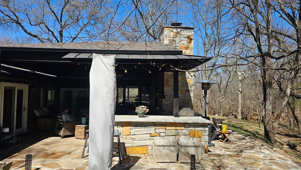 An outdoor patio with a stone fireplace, seating, and a heater is shown in a wooded area.