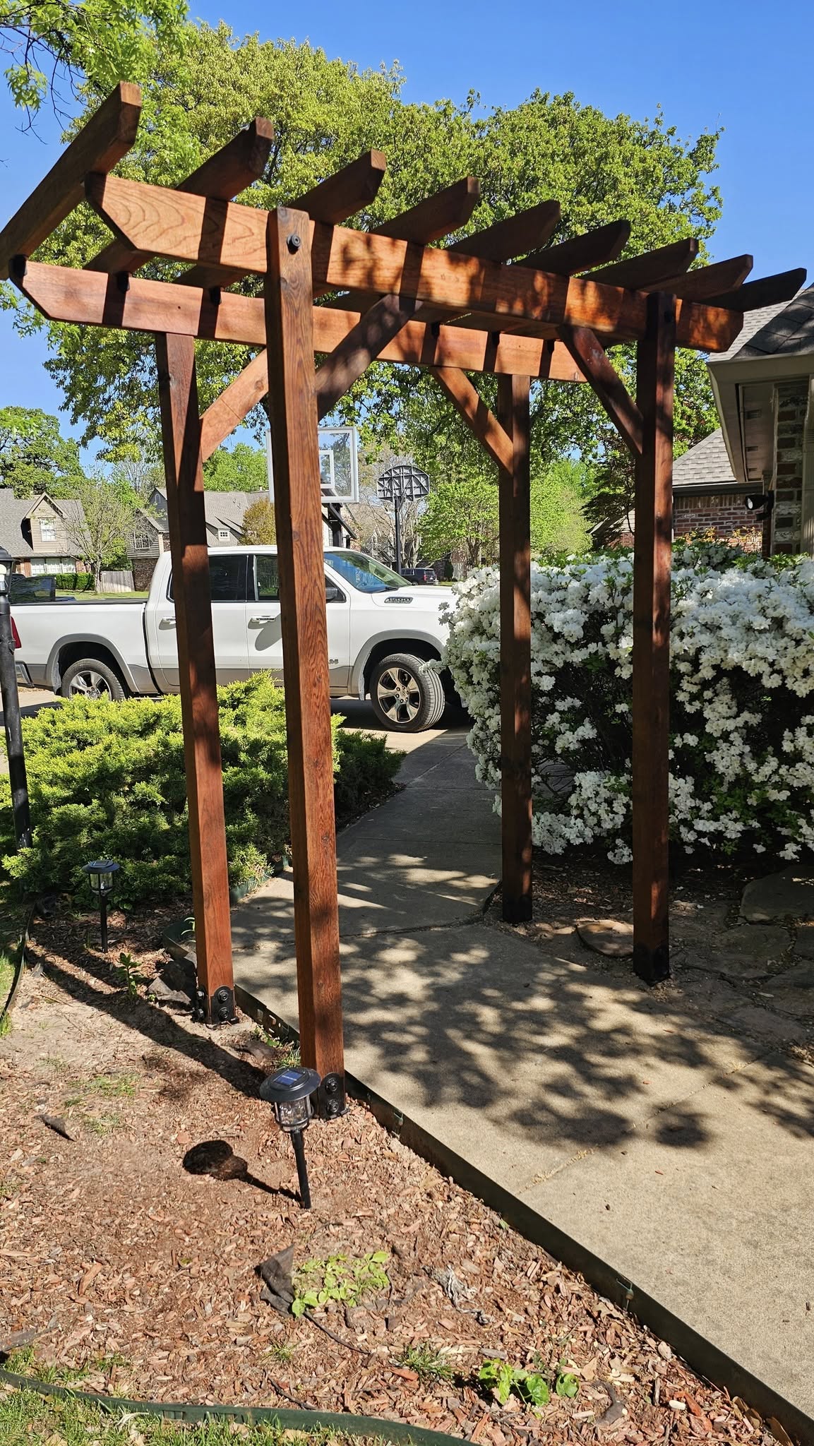 A wooden pergola stands over a walkway in a suburban yard with a white truck and flowering bushes.