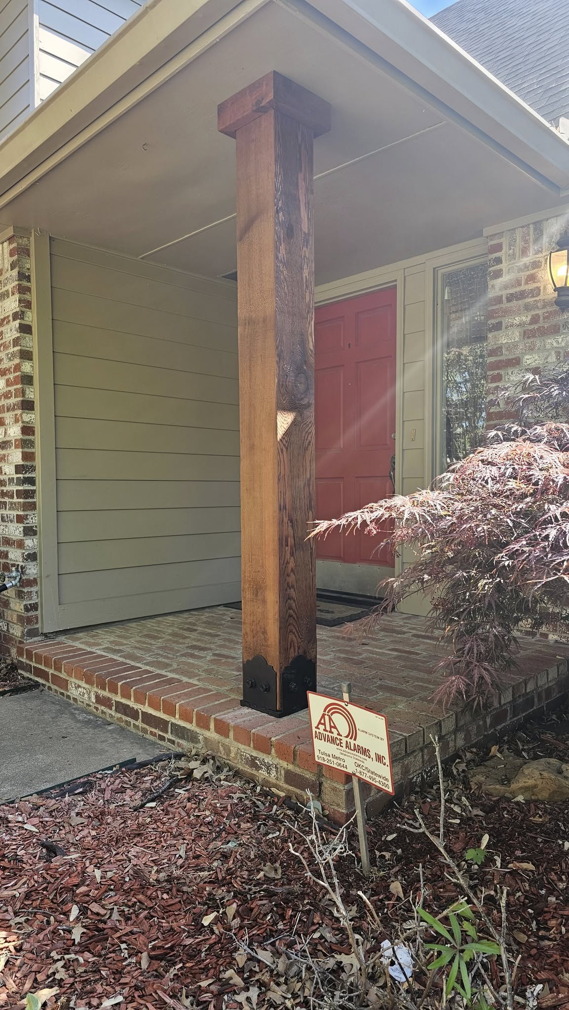 A wooden porch pillar stands in front of a red door on a brick and siding house.