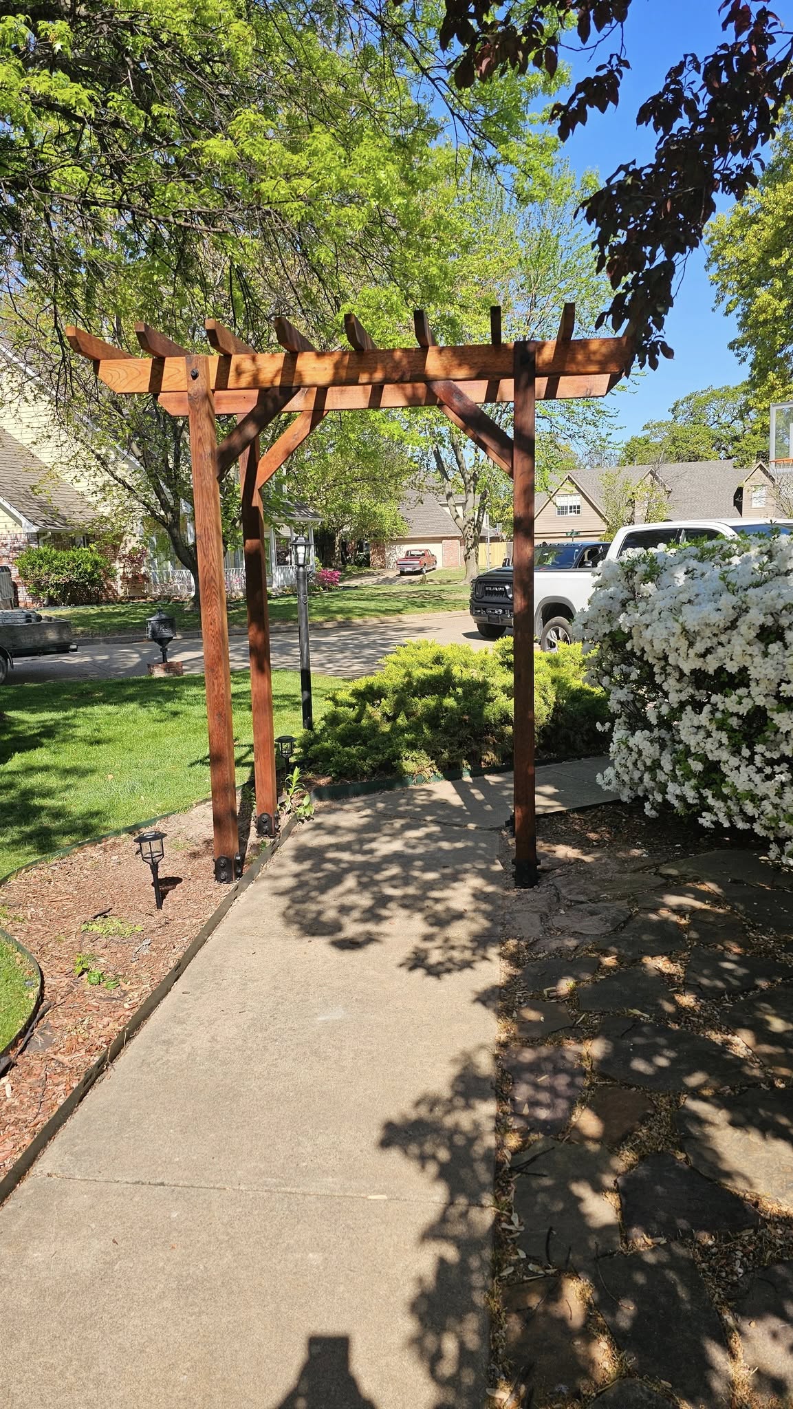 A wooden pergola stands over a concrete walkway in a suburban yard with blooming bushes and trees.