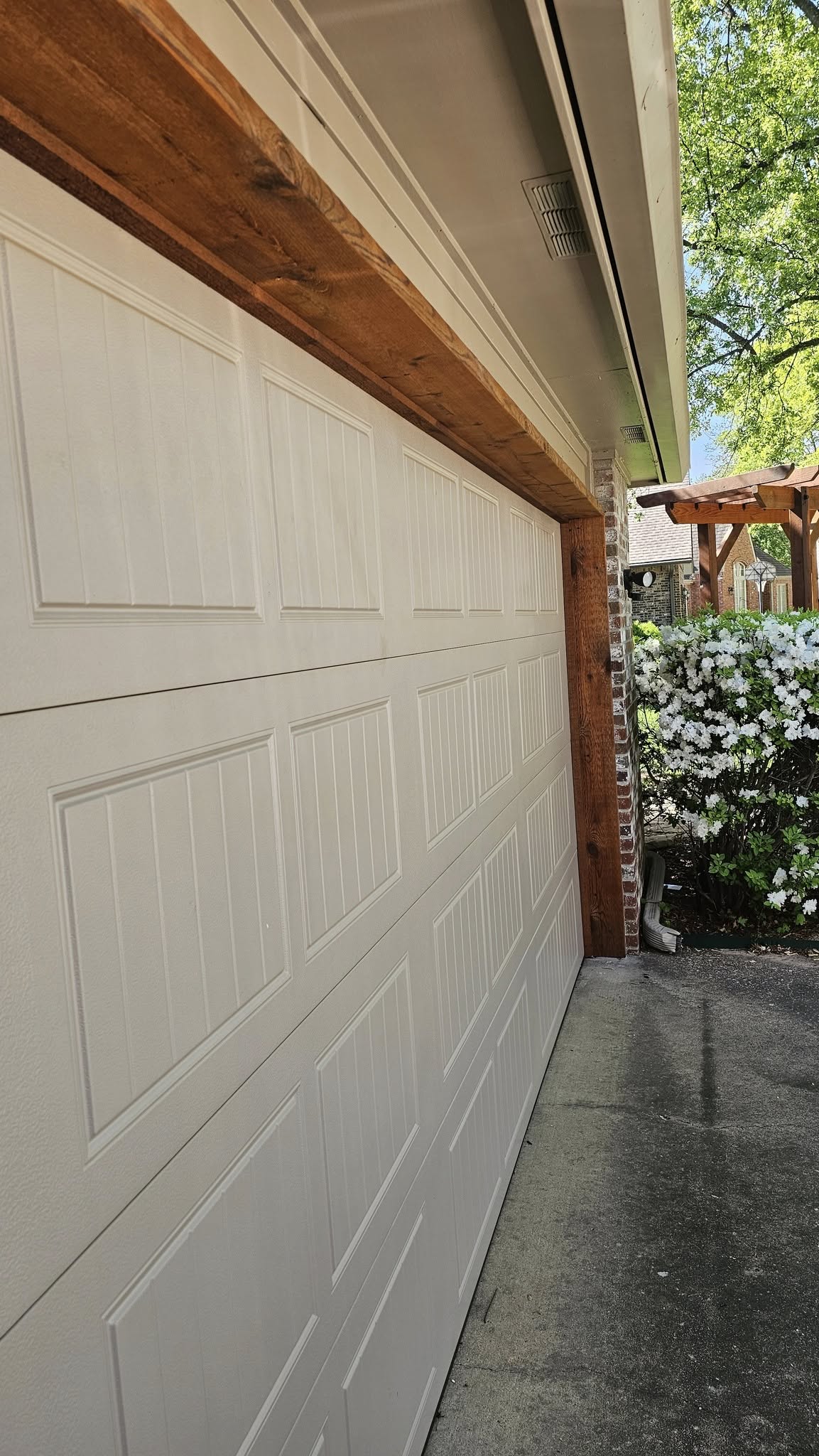 A white garage door with wooden trim is shown next to a flowering bush.