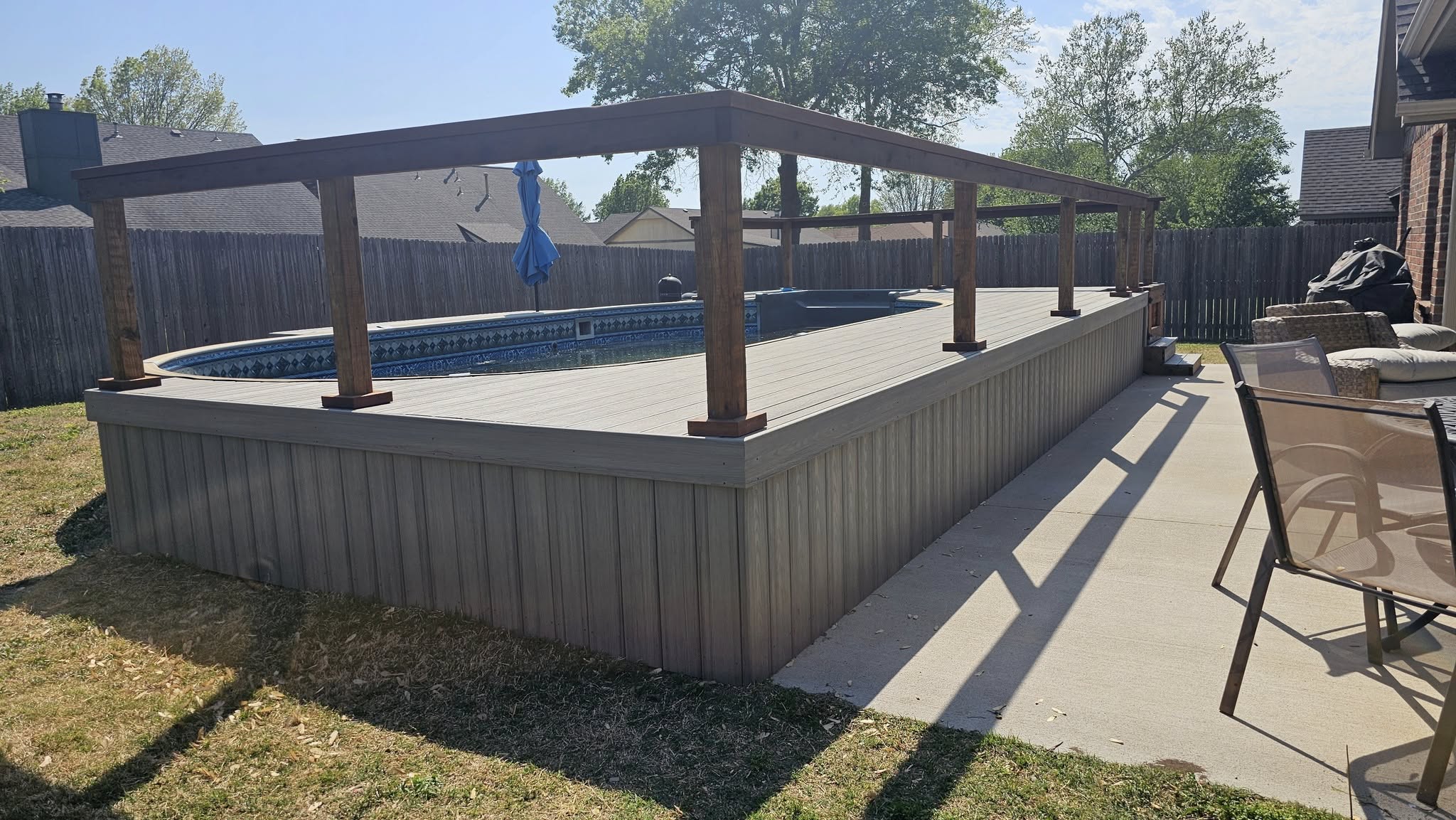 A backyard deck surrounds an oval pool with a wooden pergola overhead.