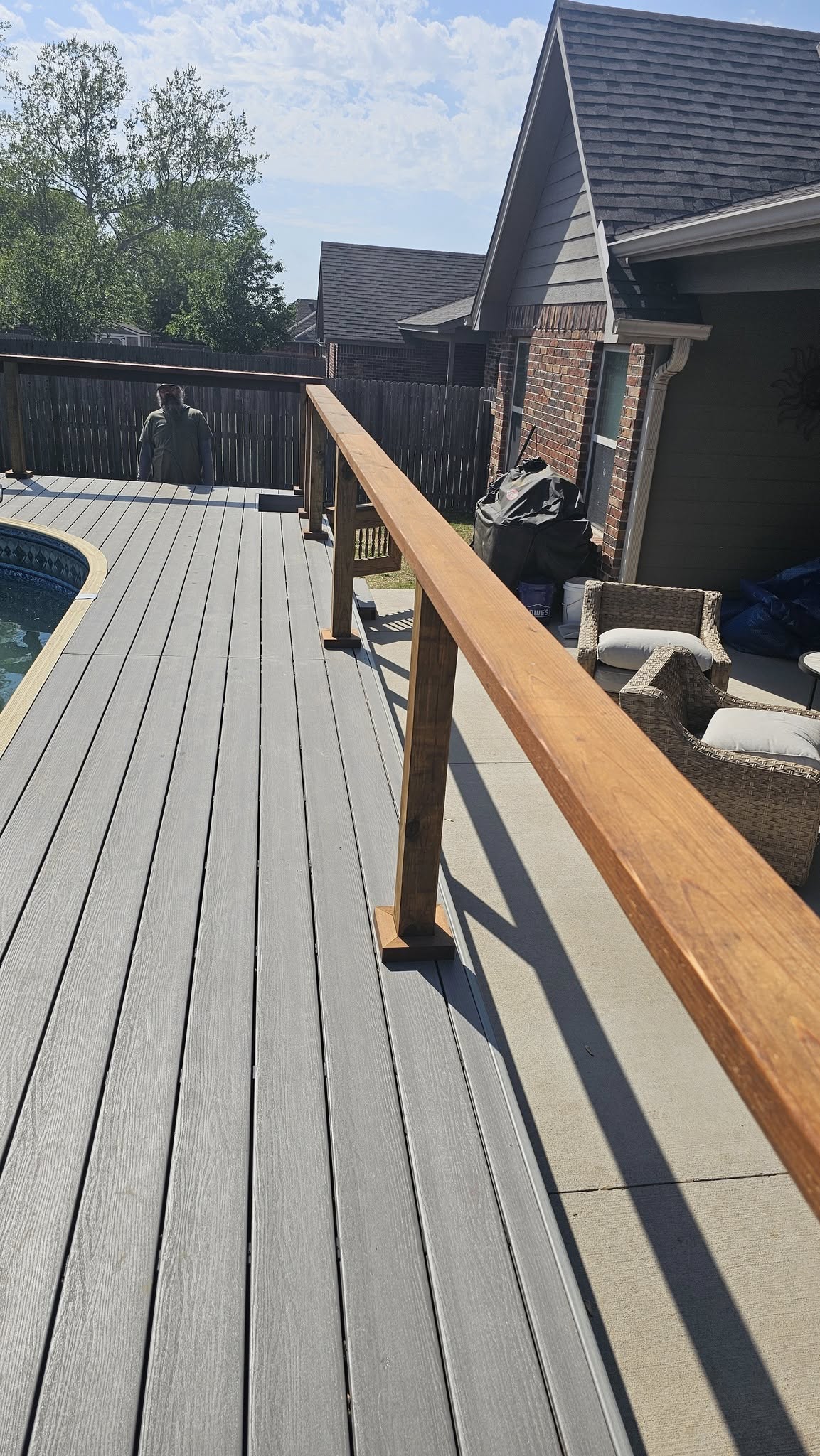 A man stands on a grey composite deck next to a pool, with a wooden railing and a house in the background.