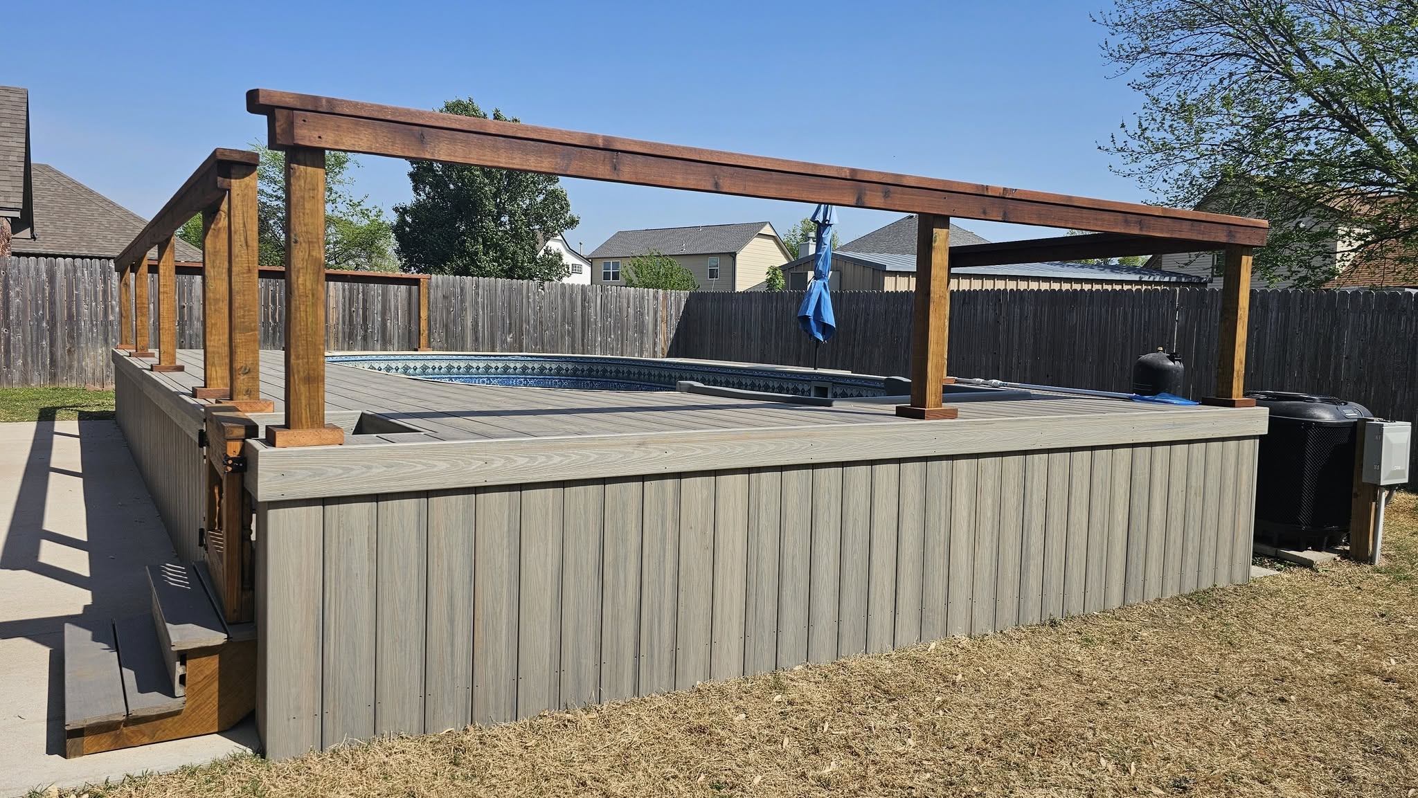 A backyard pool with a wooden deck and pergola is shown on a sunny day.