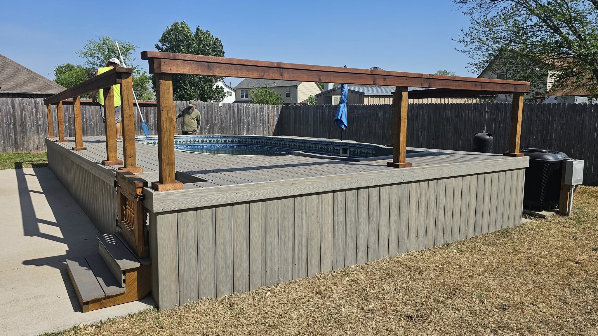 A wooden deck with a pool and pergola is shown in a backyard with two people.