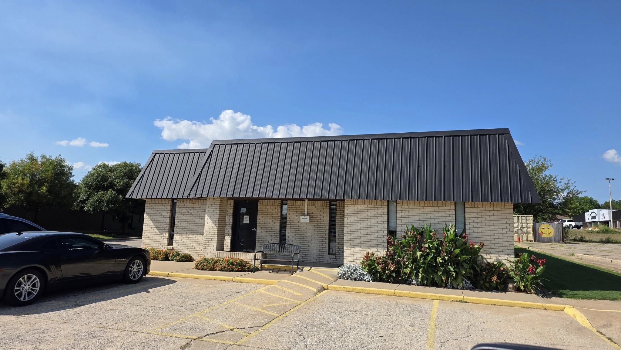 A black car is parked in front of a light brick building with a dark metal roof under a blue sky.