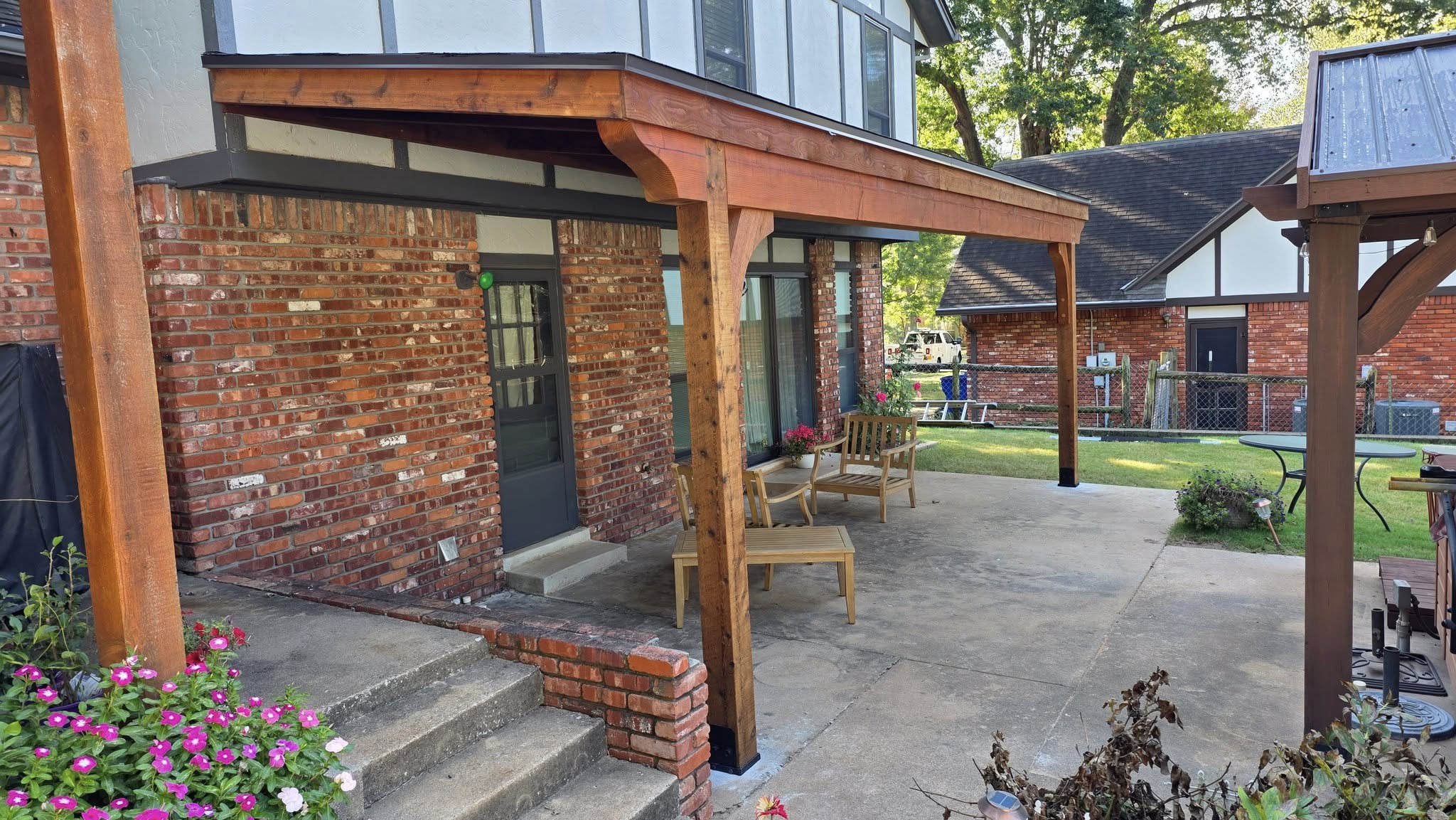 A wooden patio cover extends over a brick house with outdoor furniture and plants.
