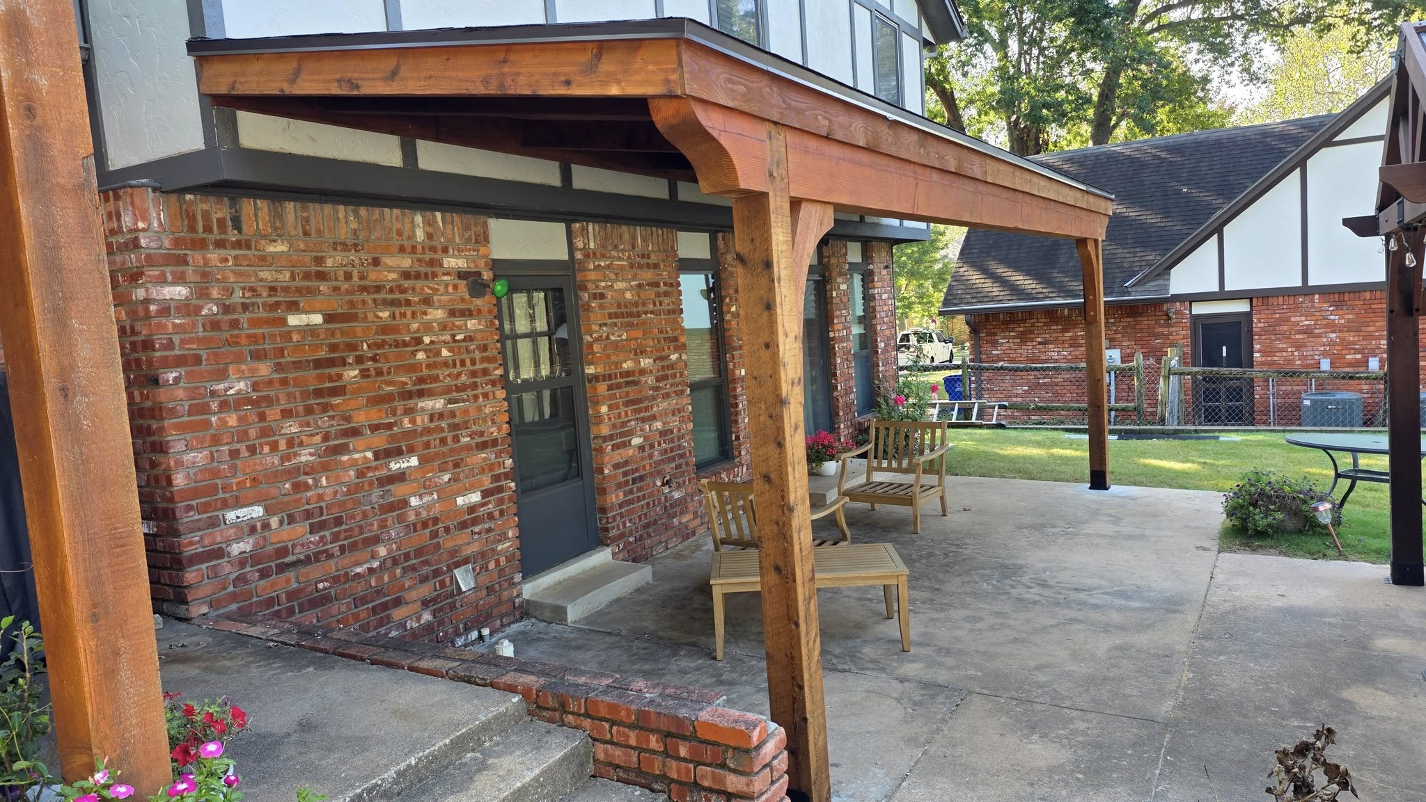 A wooden covered patio with furniture is shown outside a brick house.