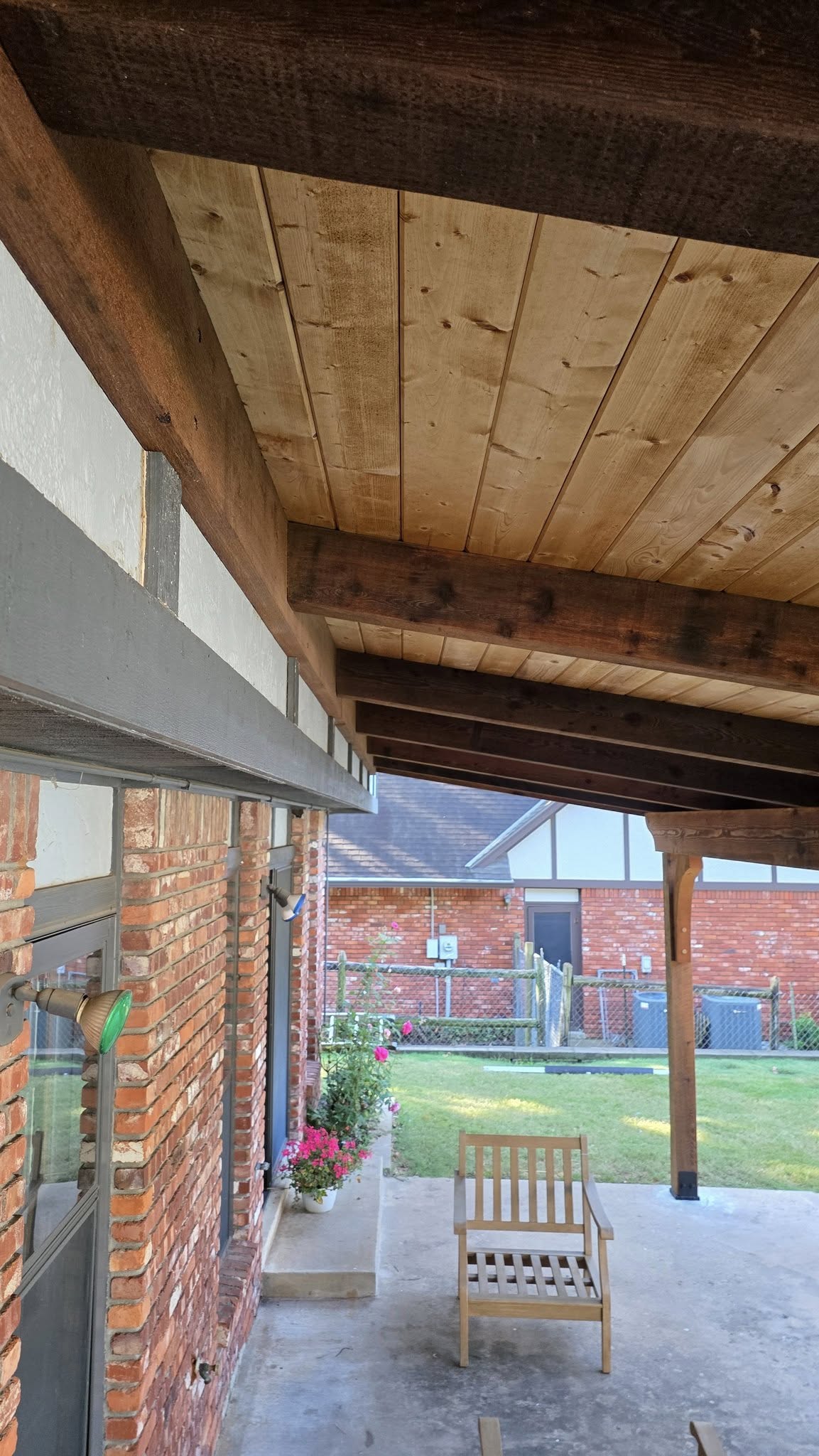A wooden chair sits on a patio under a wooden-beamed roof, next to a brick house.