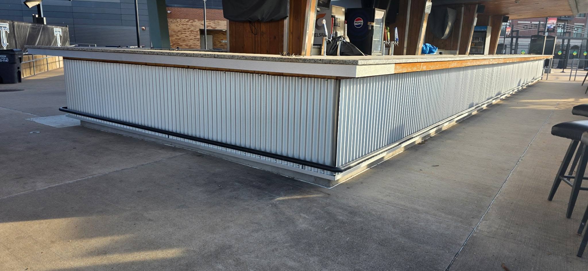 An outdoor bar with corrugated metal siding and stools is shown on a concrete patio.