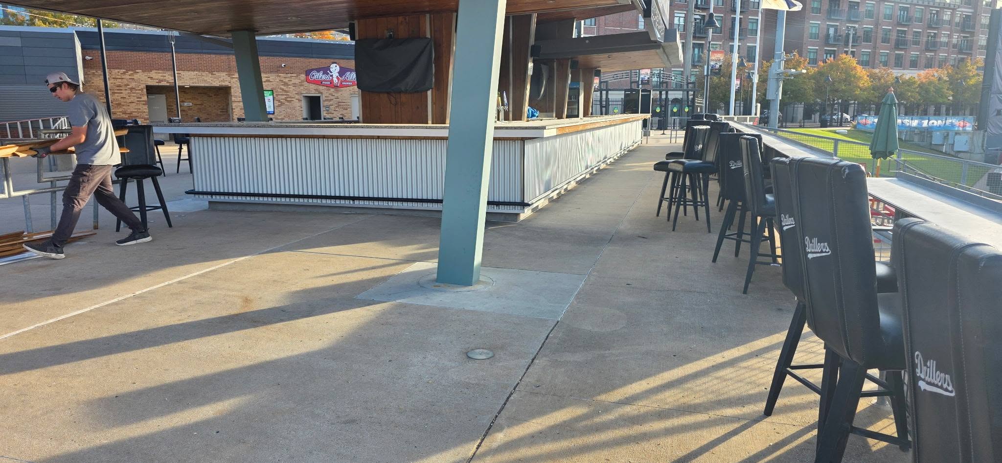 A man carries wood near a bar with stools outside a building.