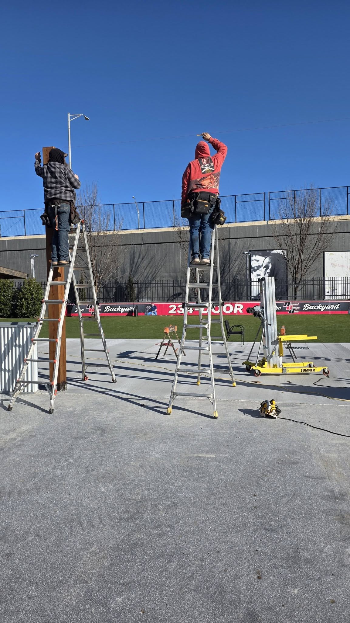 Two construction workers on ladders install a sign in an outdoor area with a stadium in the background.