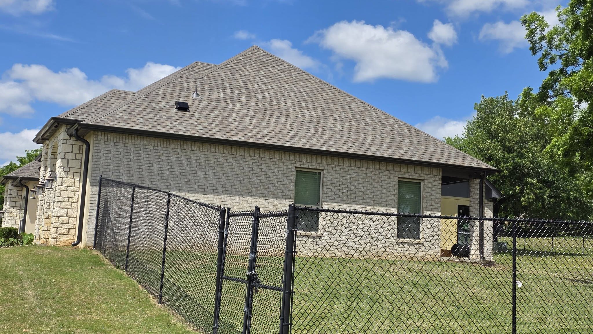 A chain-link fence encloses a brick house with a shingled roof under a blue, cloudy sky.
